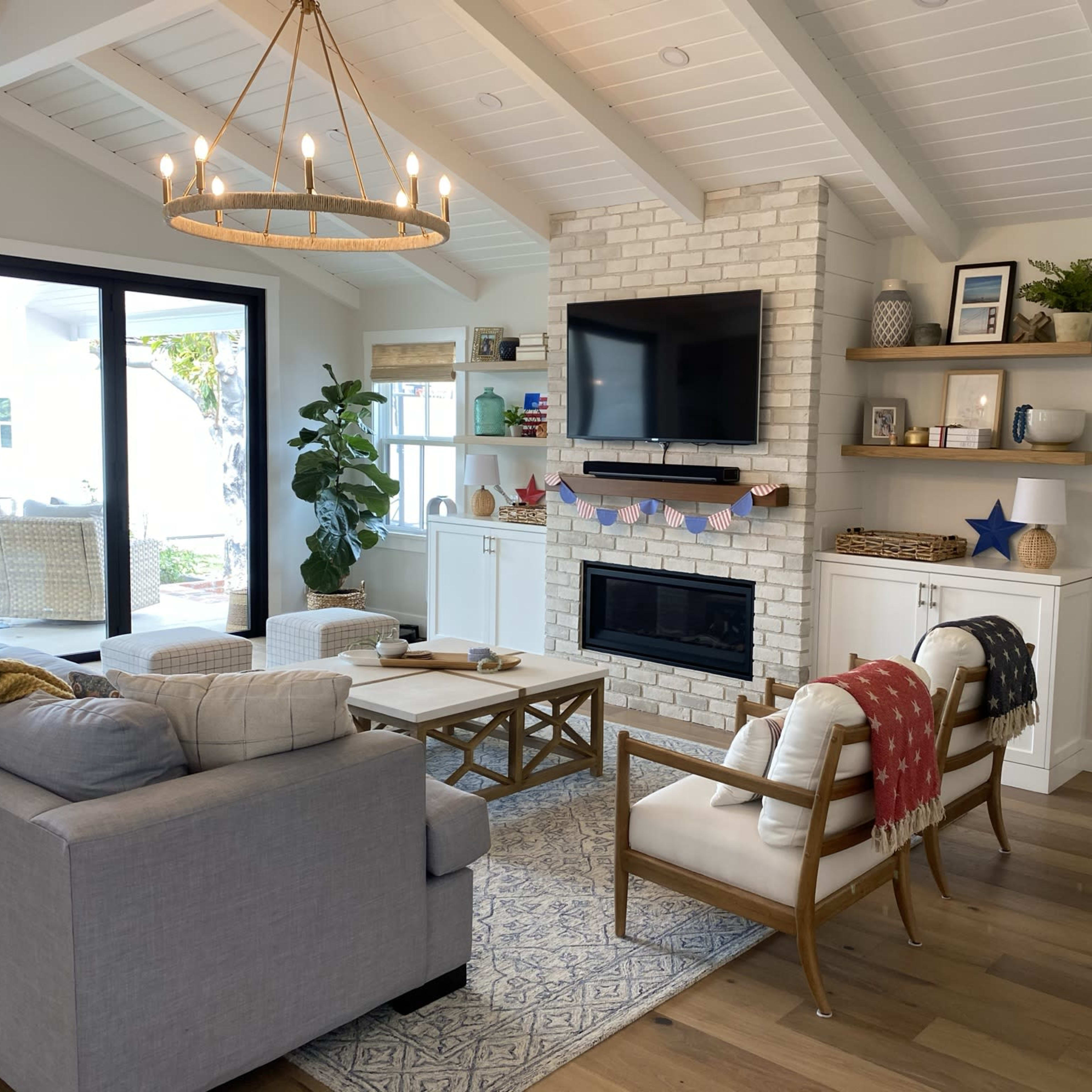 The living room features a gray sofa, wooden coffee table, and a white brick fireplace, with large windows allowing natural light and a view of greenery outside.