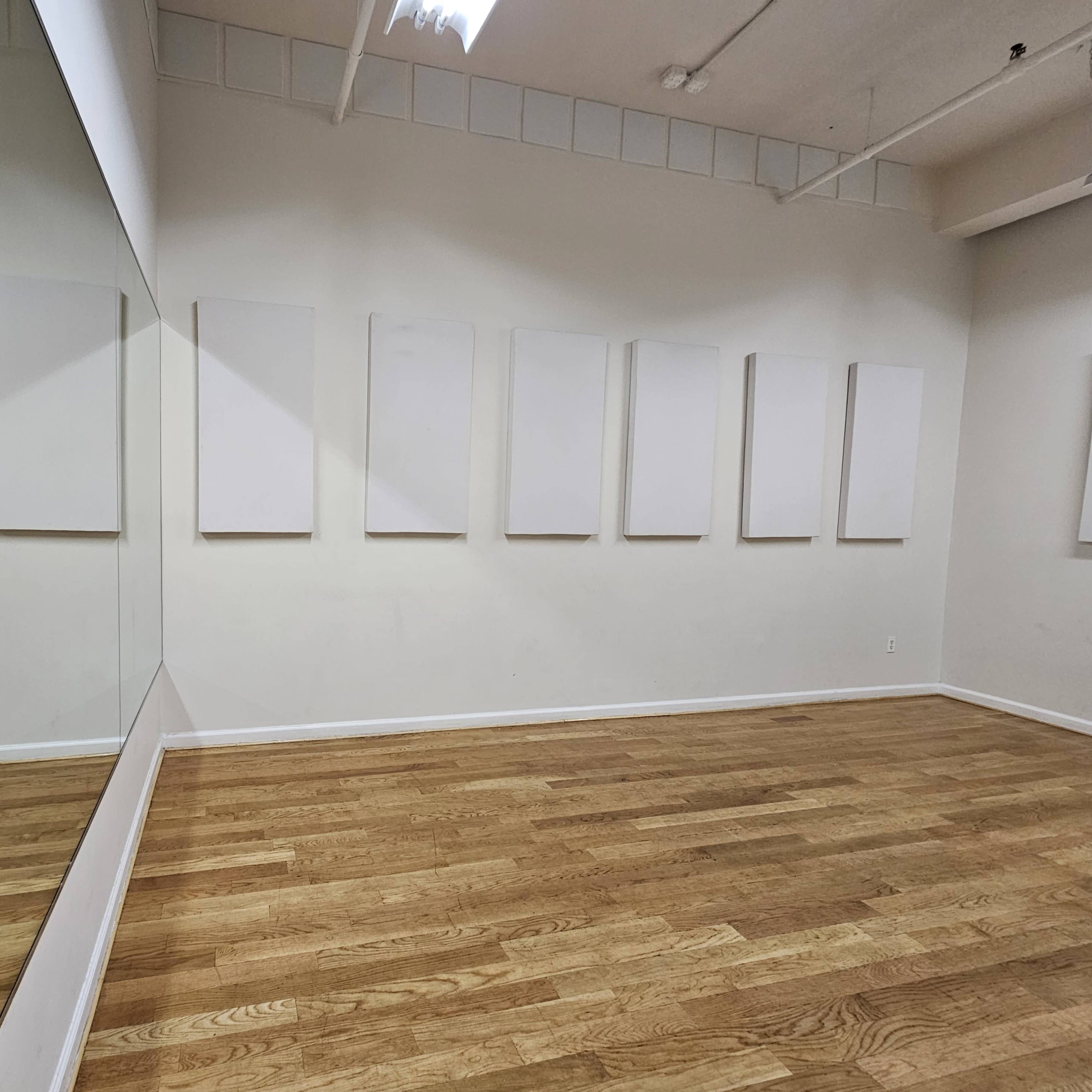 An empty room with light wooden flooring, white walls, and a large mirror on one side reflecting blank canvases arranged on the opposite wall.