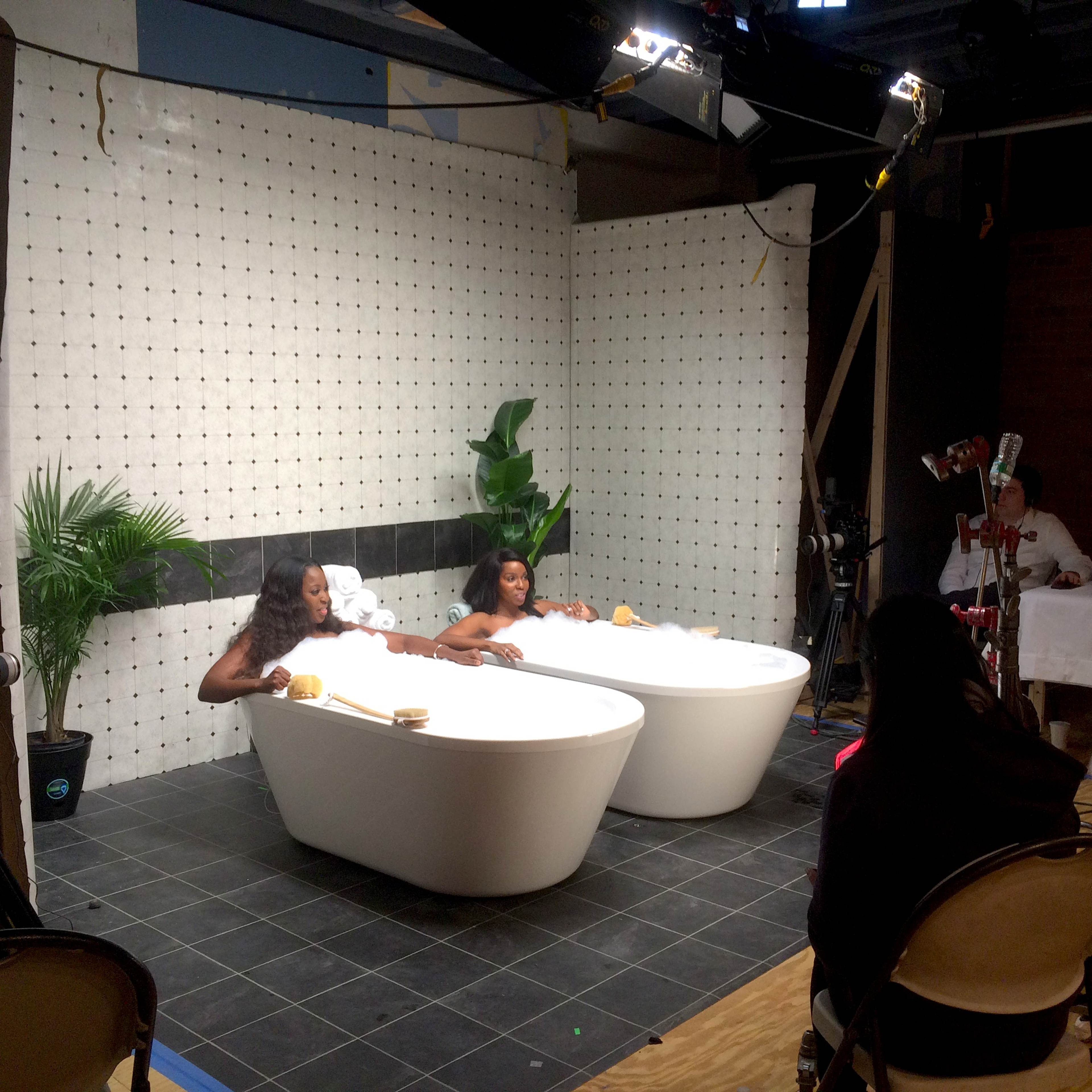 Two models are reclining in separate bathtubs on a set designed for a photoshoot, while a small crew prepares to capture the scene.