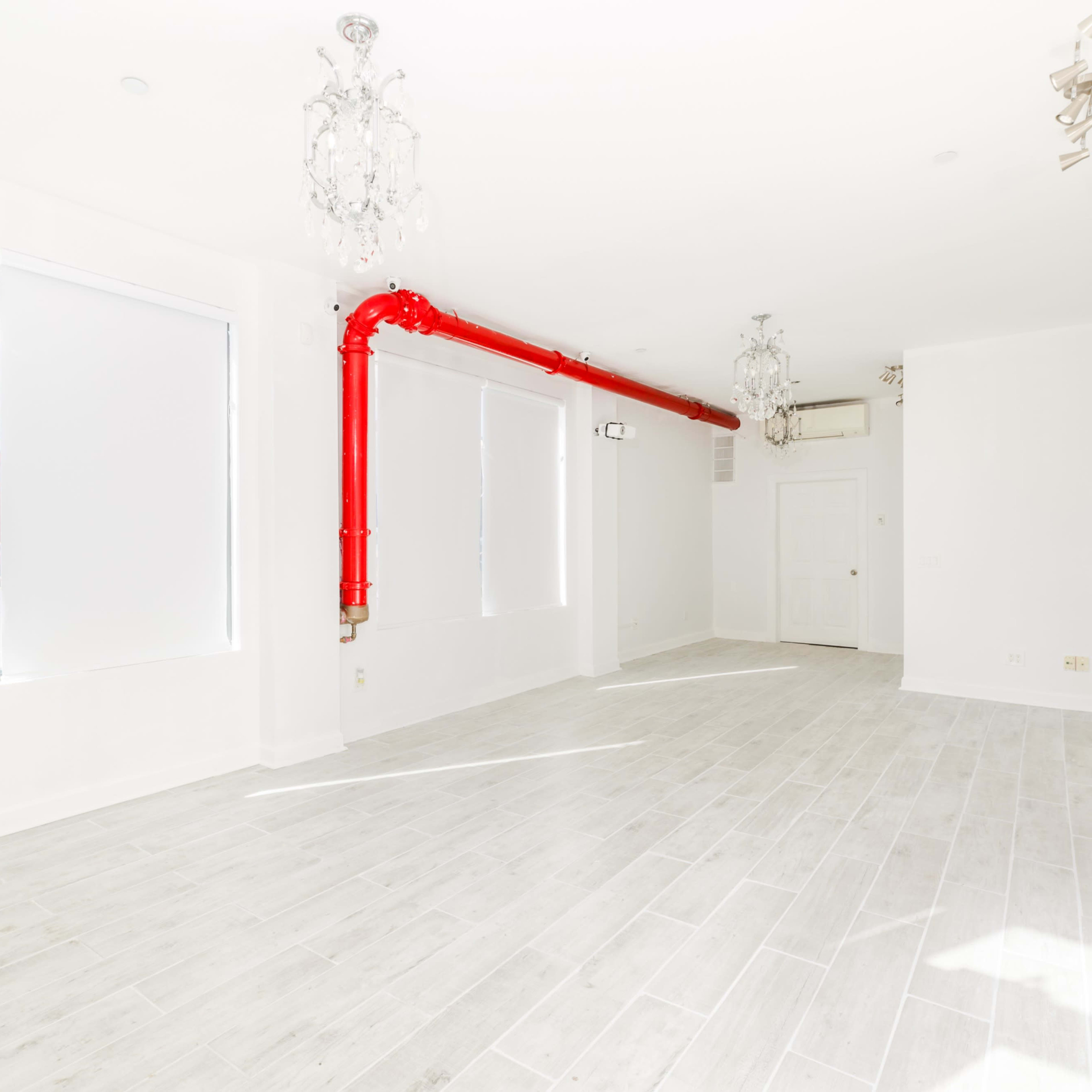 A spacious, empty room with light-colored flooring, large windows with covered blinds, and a prominent red pipe running along the ceiling.
