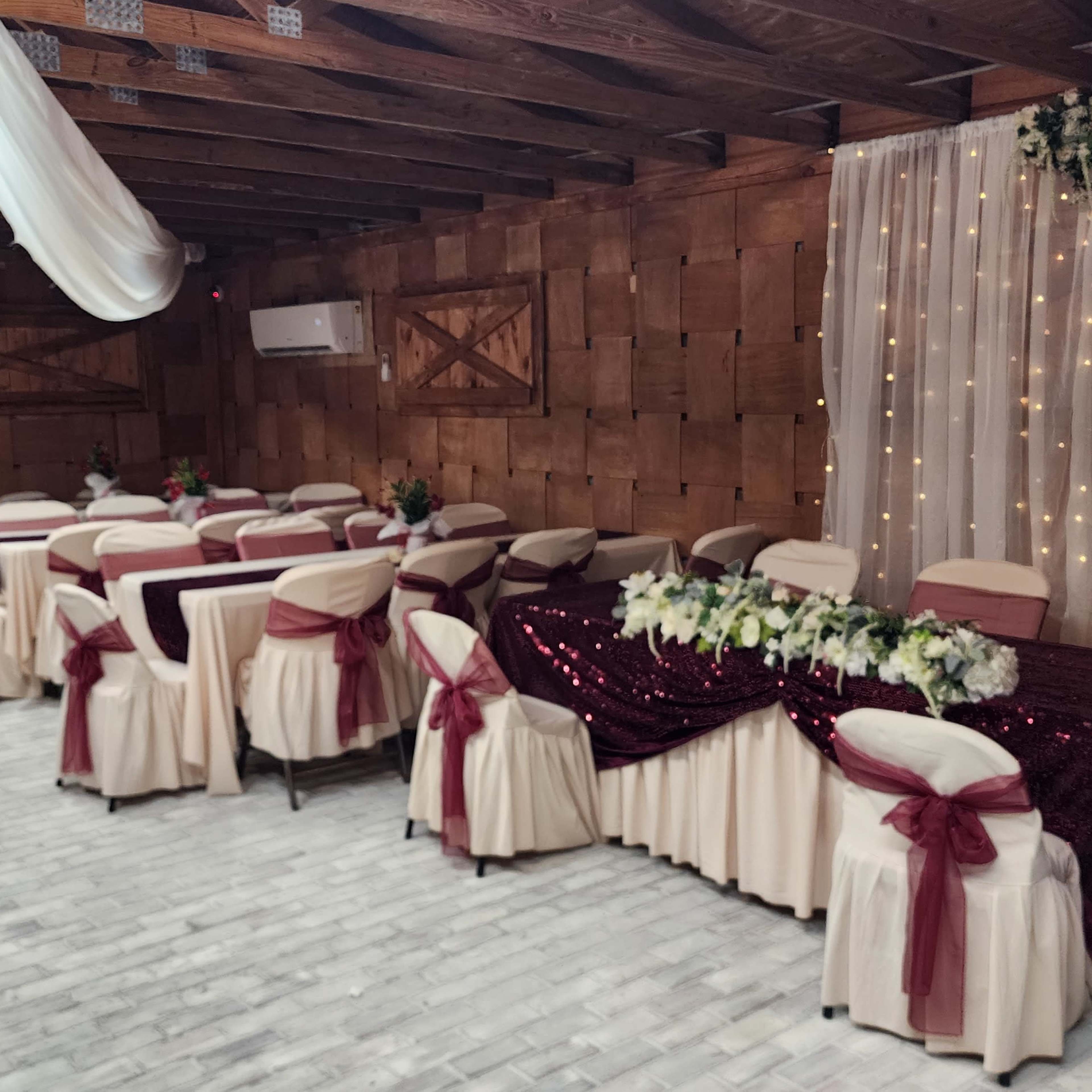 A decorated indoor space with several tables arranged for an event, featuring white tablecloths with maroon sashes and a backdrop of draped fabric with decorative lights and floral arrangements.
