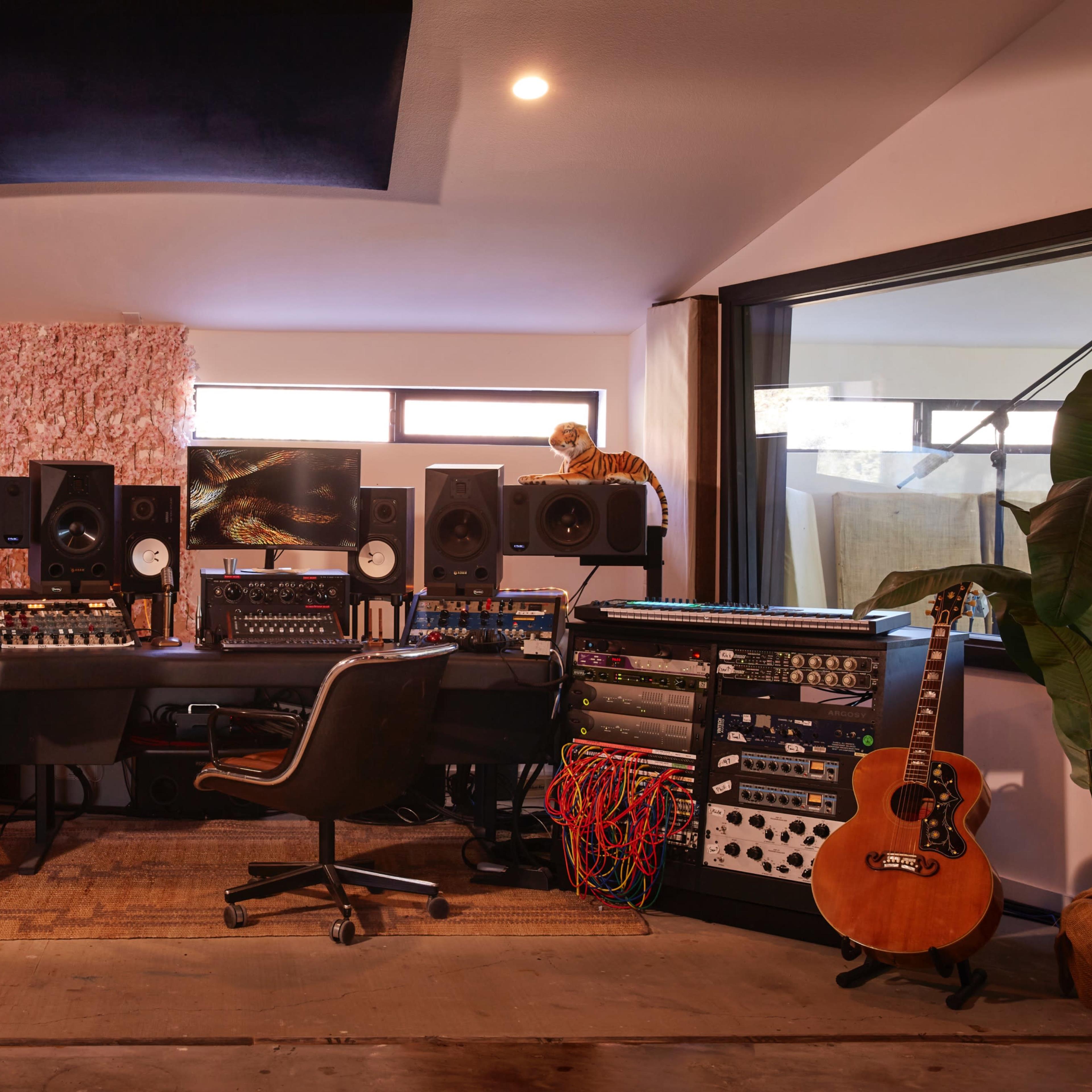 A music recording studio is shown, featuring a central desk with audio equipment, speakers, and a vintage guitar, surrounded by various recording and mixing devices.