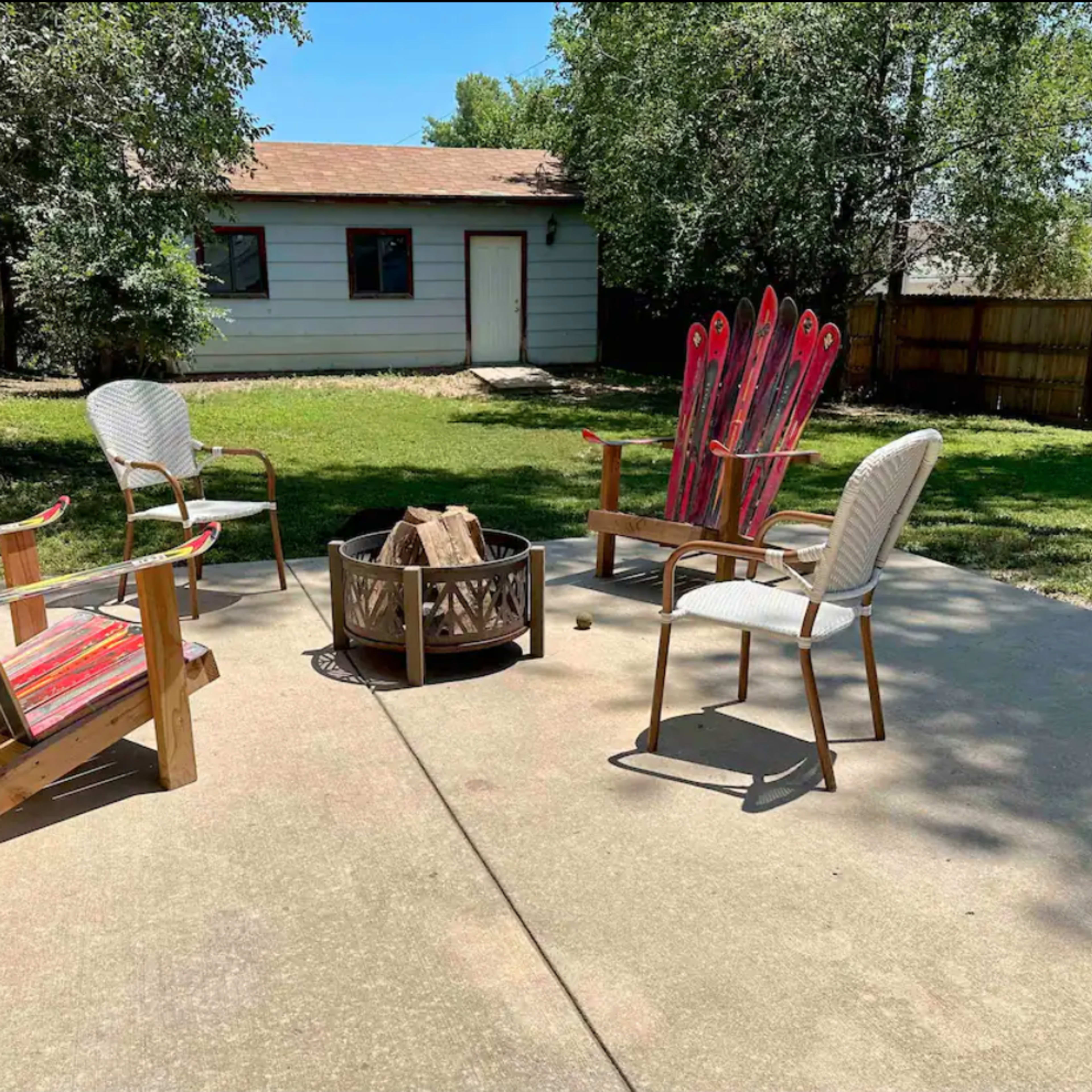 A patio area features four chairs surrounding a fire pit with a wood pile, set against a backdrop of a house and green trees.