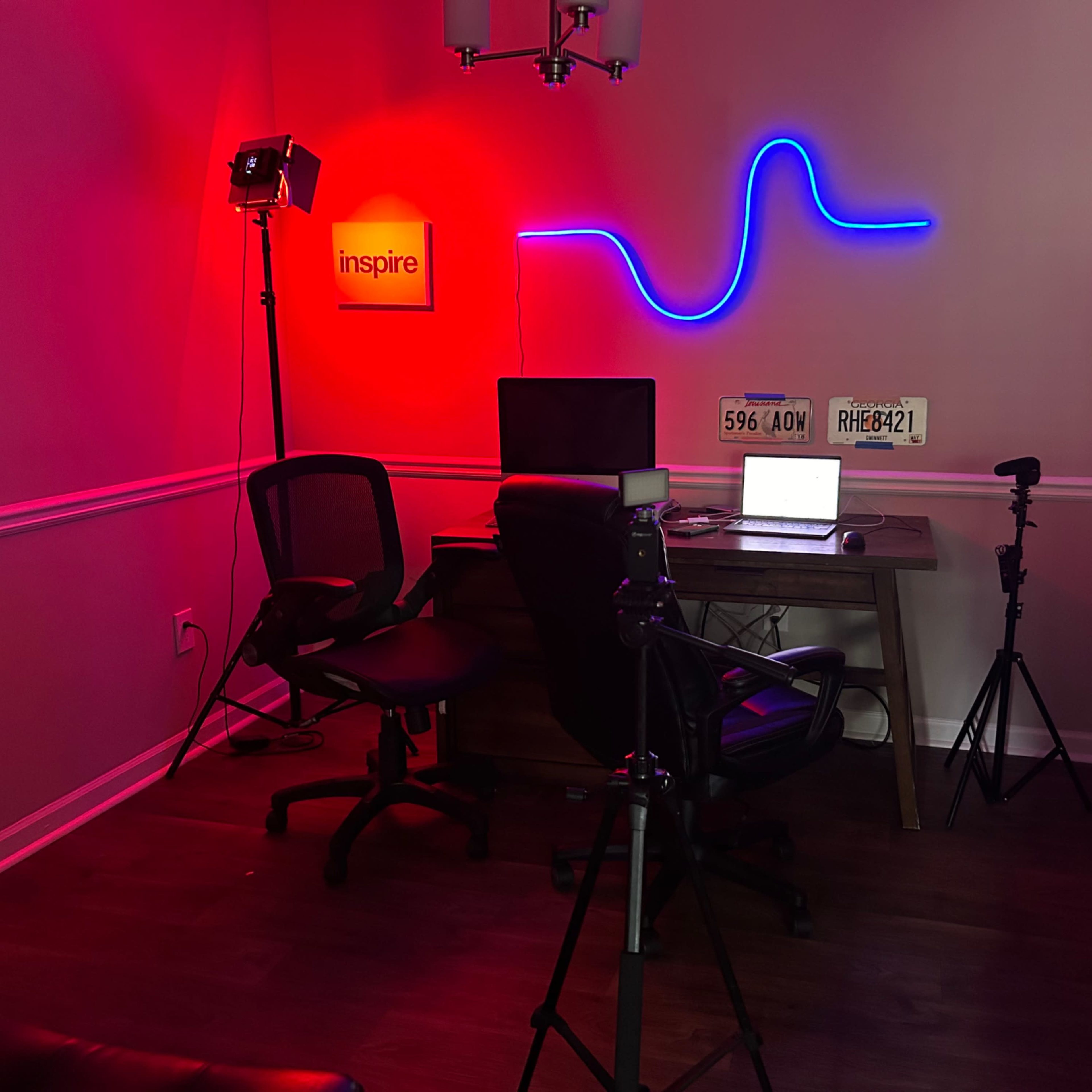 A home office setup features a desk with a computer, two chairs, a neon light display, and various filming equipment.