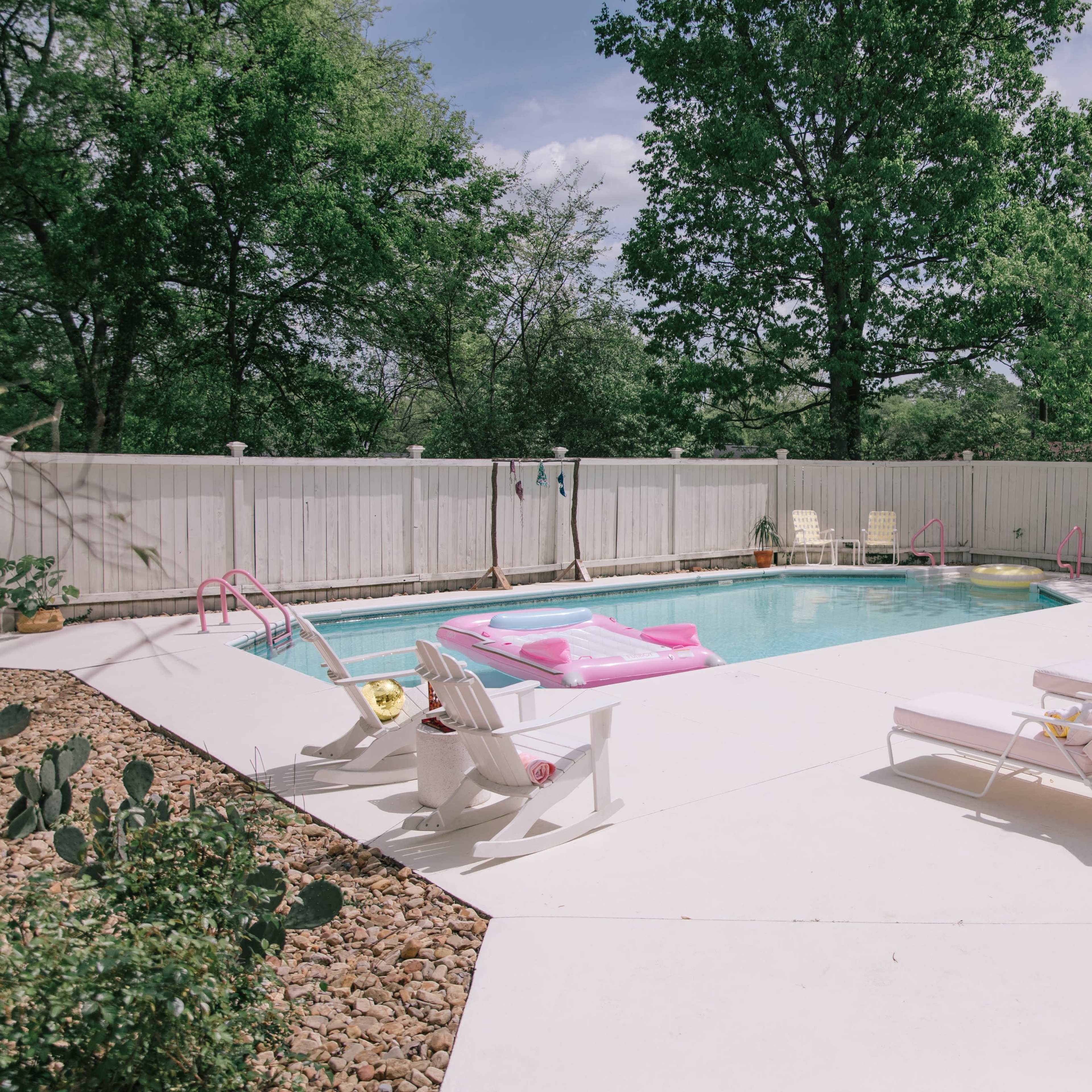 A backyard pool area with several lounge chairs, pink floats in the water, and tall trees and a white fence surrounding the space.