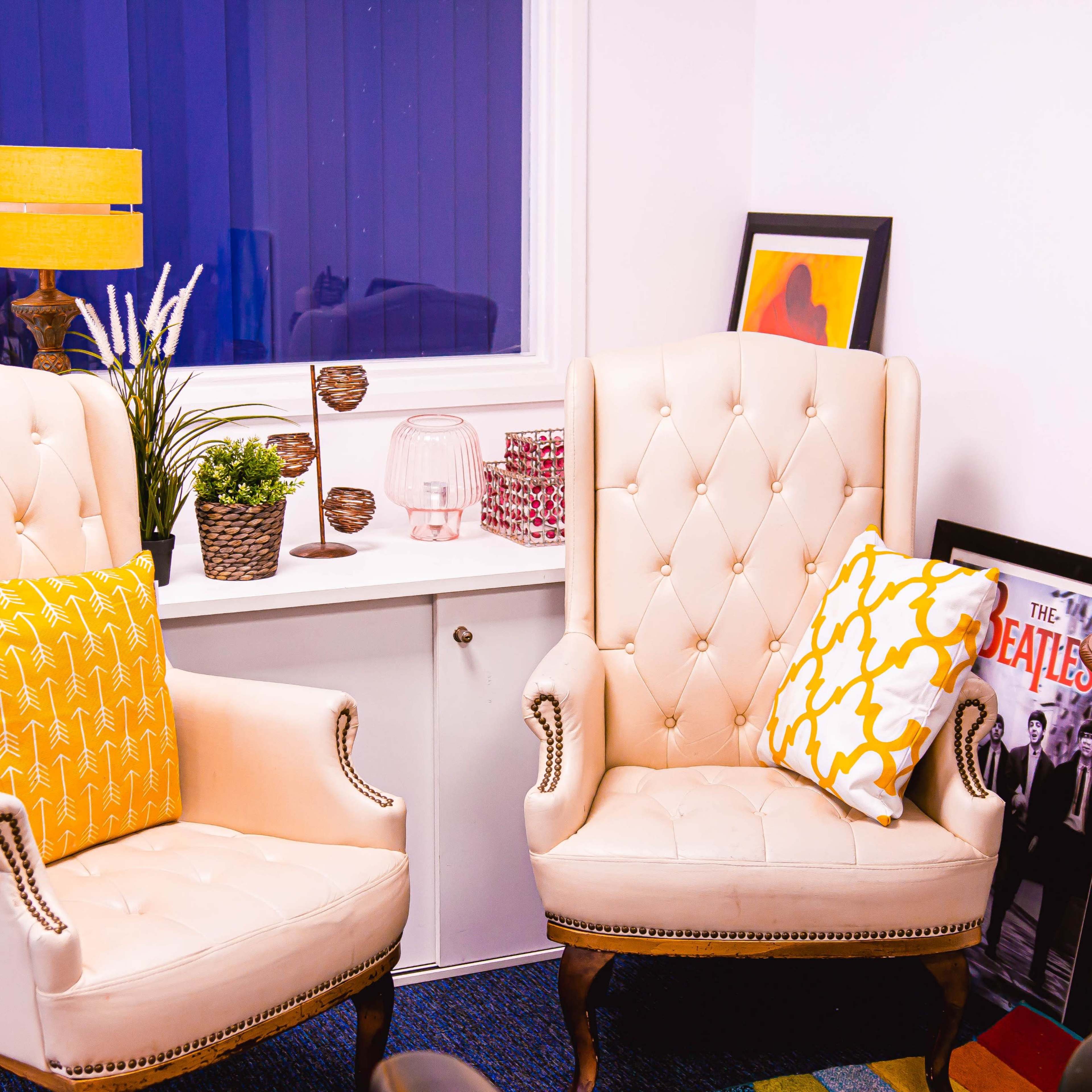 Two cream-colored upholstered armchairs with decorative pillows, positioned next to a small white table against a wall with artwork and a lamp.