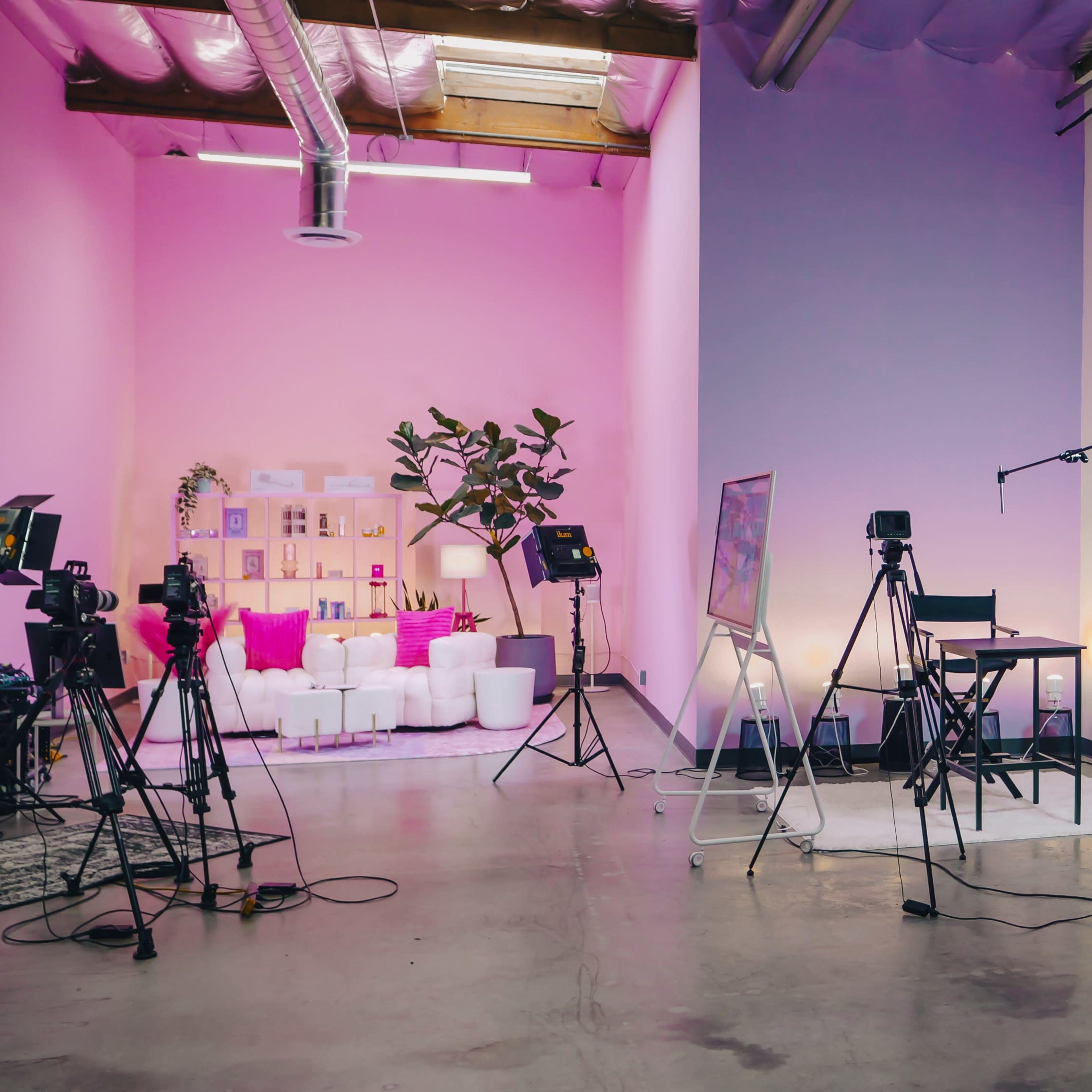A studio space features various camera setups and lighting equipment against a backdrop of pink and purple walls.