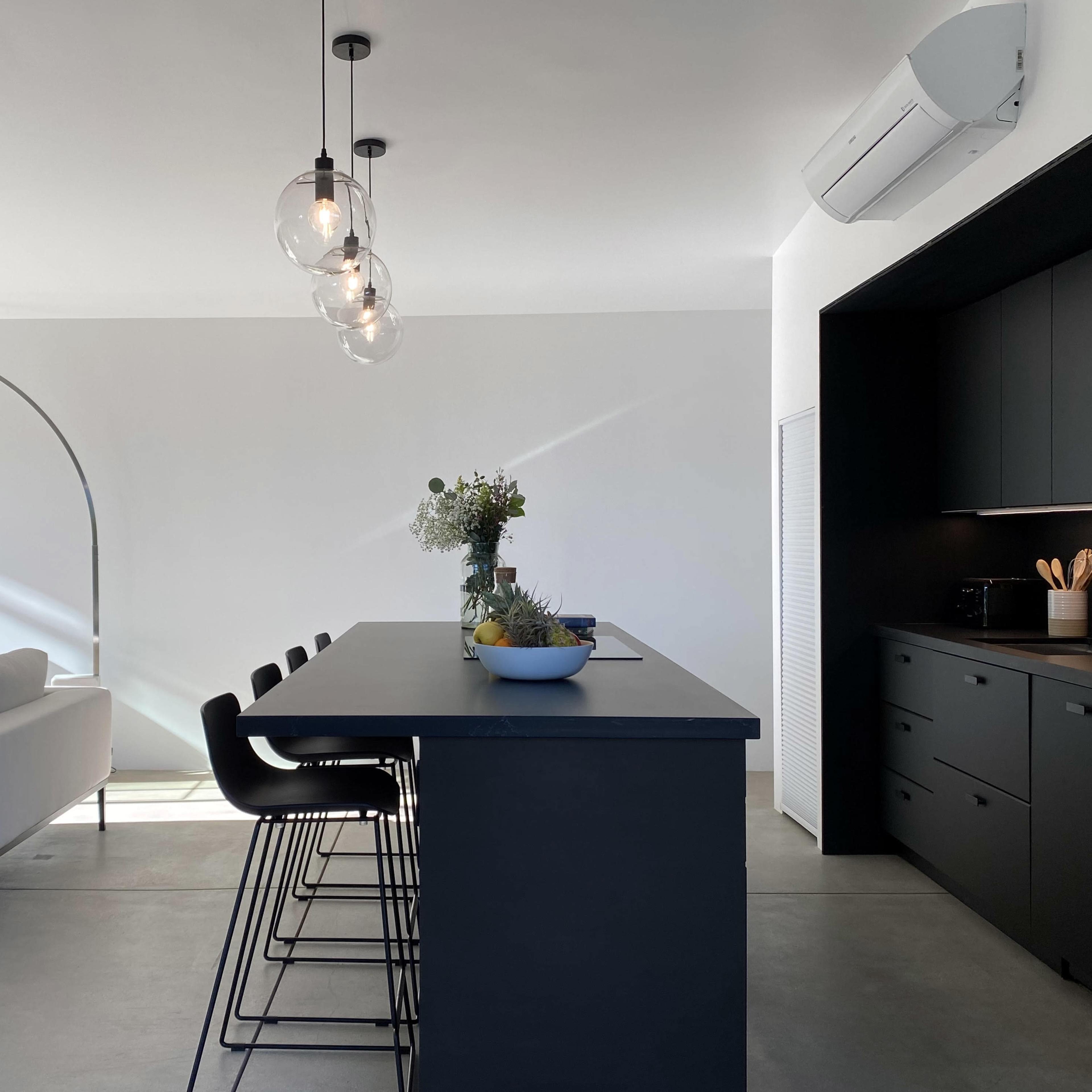 A modern kitchen with a black island bar, minimalist decor, and large windows allowing natural light to enter the space.