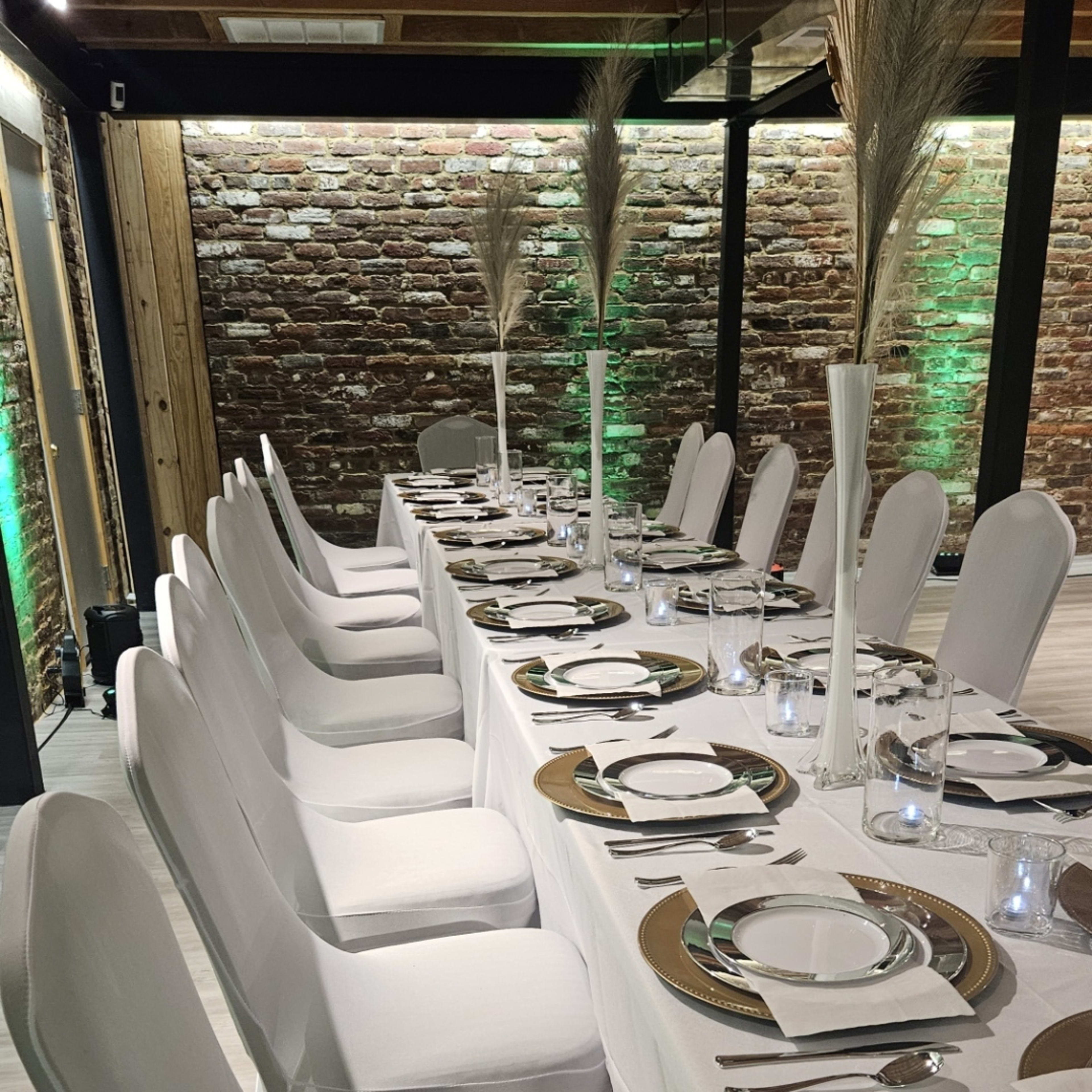 A long dining table set with white tablecloths and chairs, featuring plates, glasses, and decorative centerpieces, is arranged in a room with exposed brick walls and green lighting.