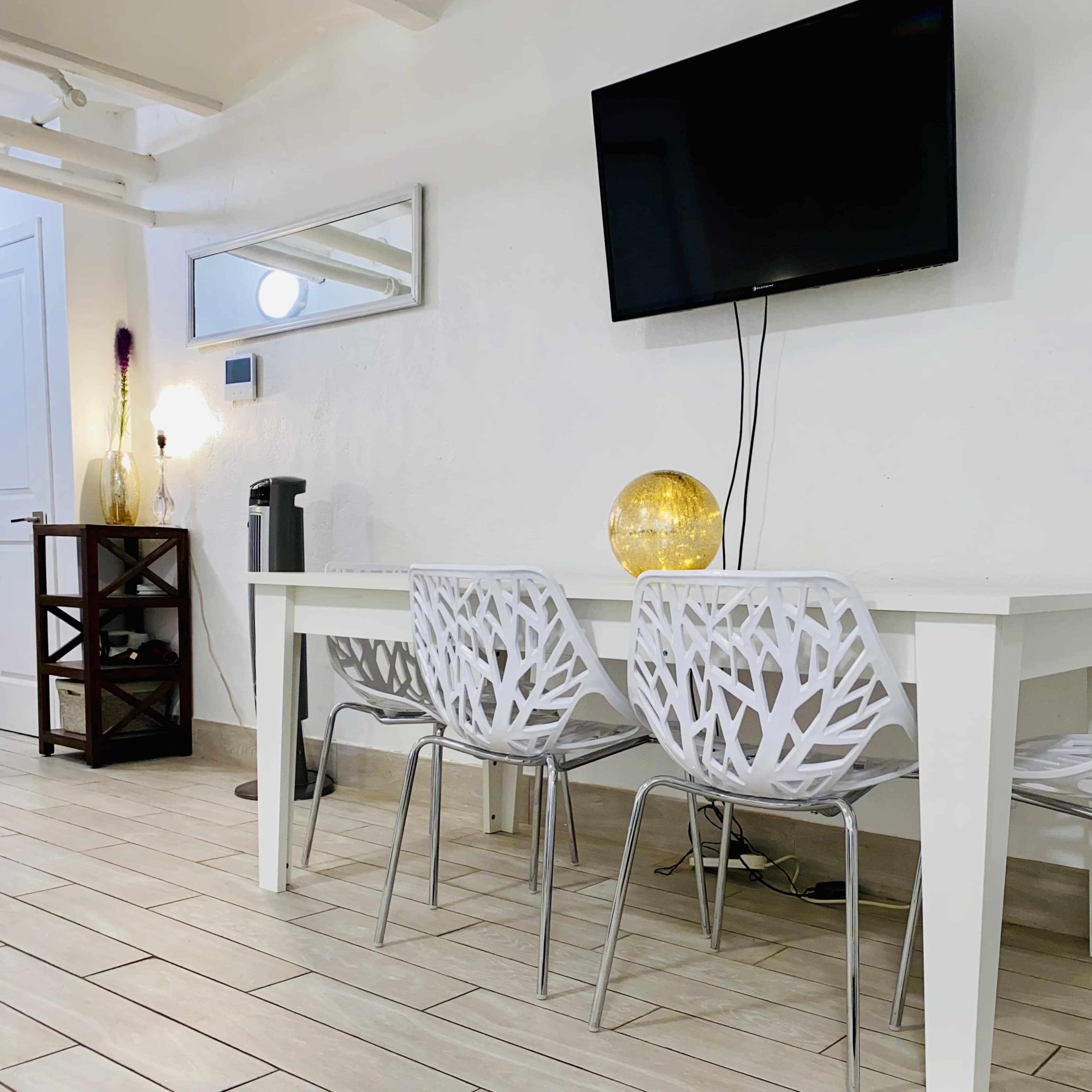 A brightly lit room features a white dining table with three patterned chairs, a wall-mounted television, and a decorative lamp.
