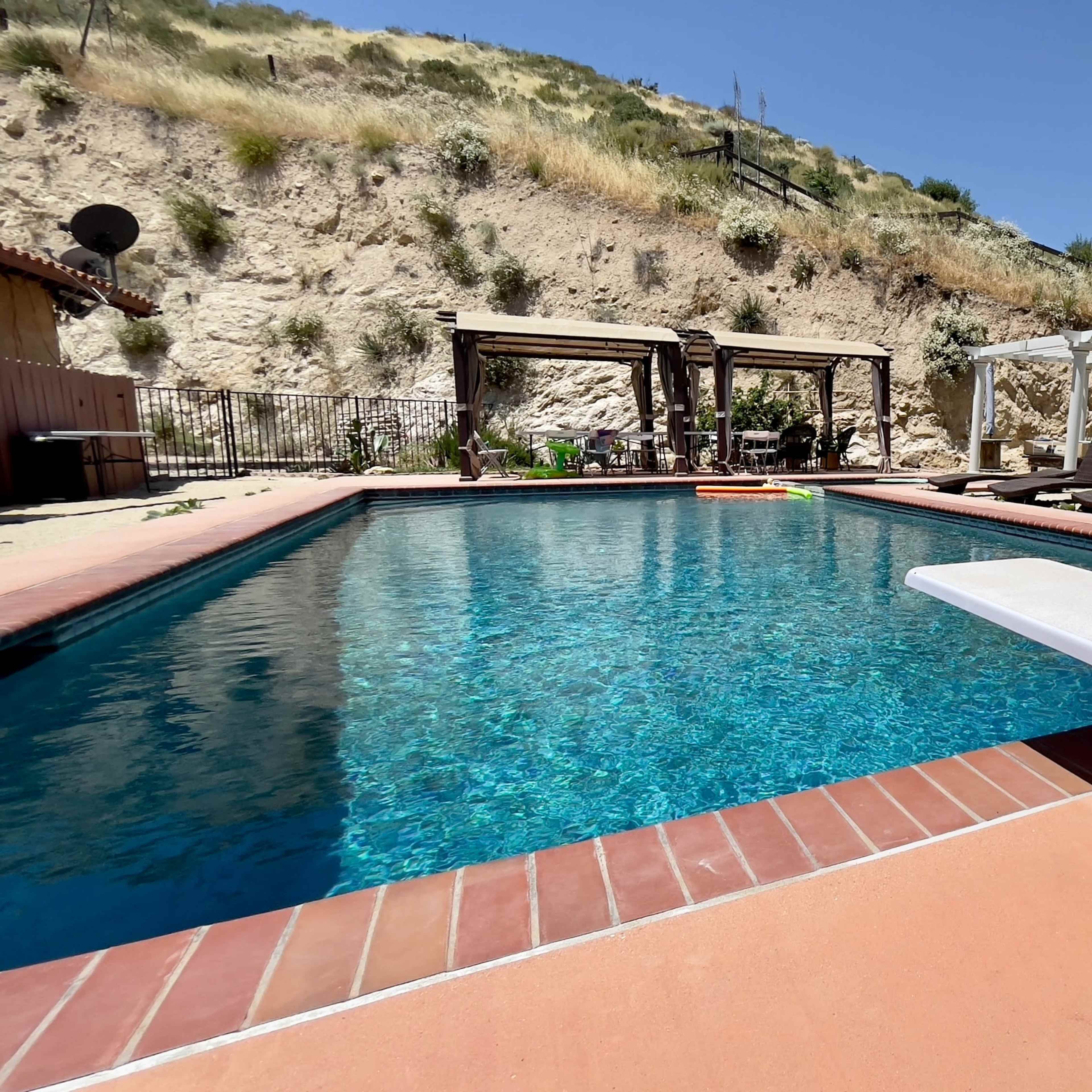 A swimming pool with a diving board, surrounded by lounge chairs and shaded seating areas, set against a hillside backdrop.