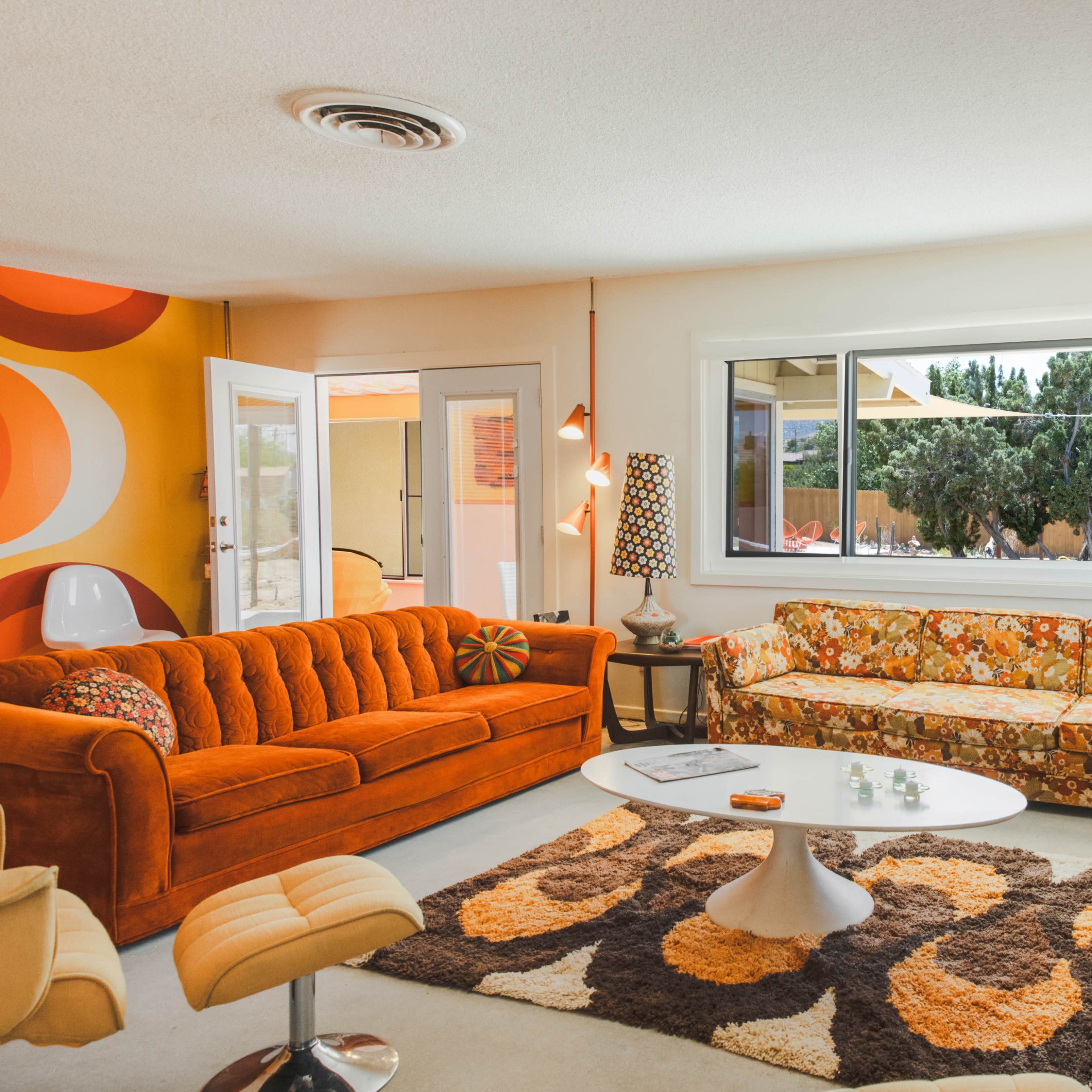 The living room features a vibrant color scheme with an orange velvet sofa, a floral-patterned couch, a round white coffee table, and a retro egg chair.