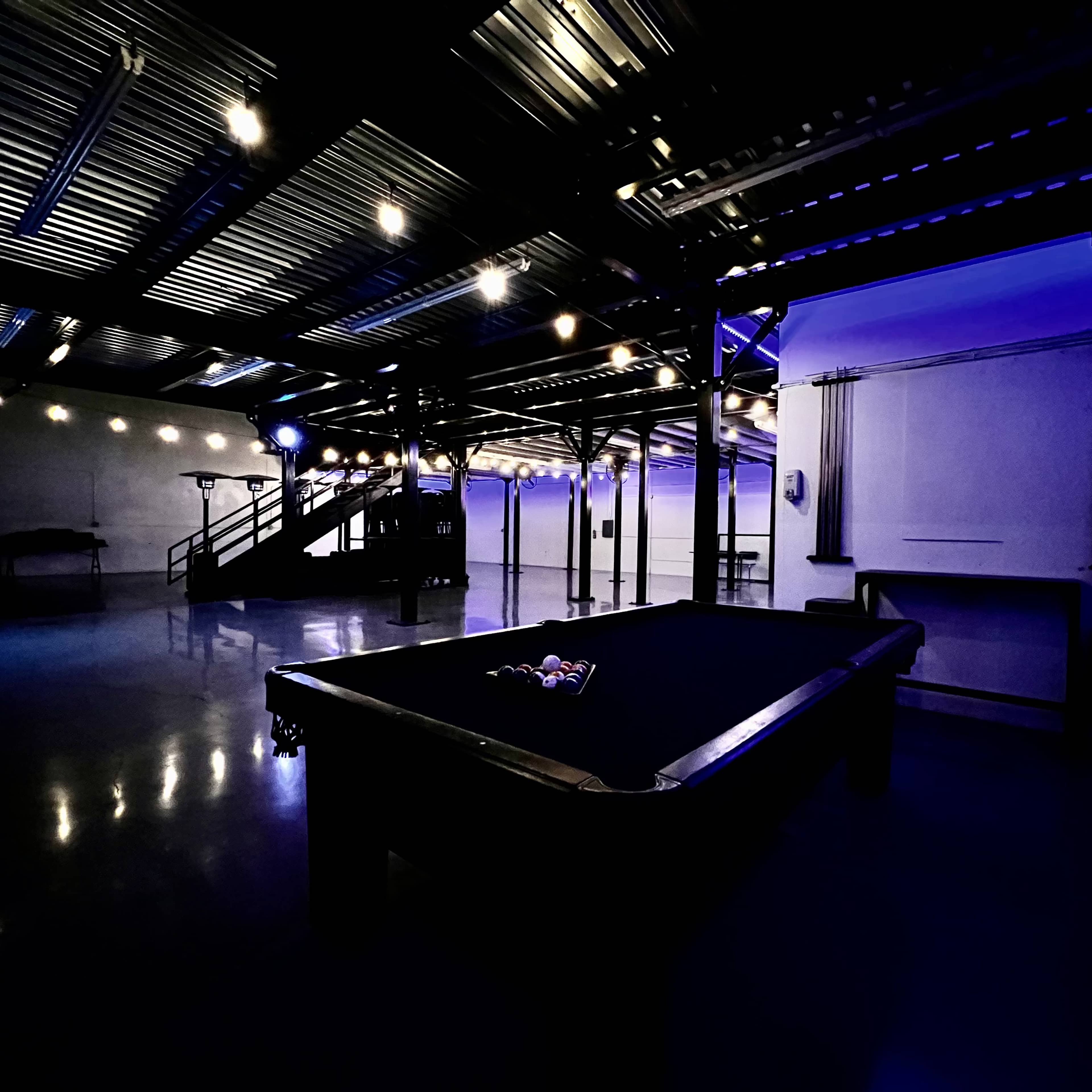 A dimly lit room with a billiard table in the foreground and a staircase leading to an upper level in the background, illuminated by string lights.