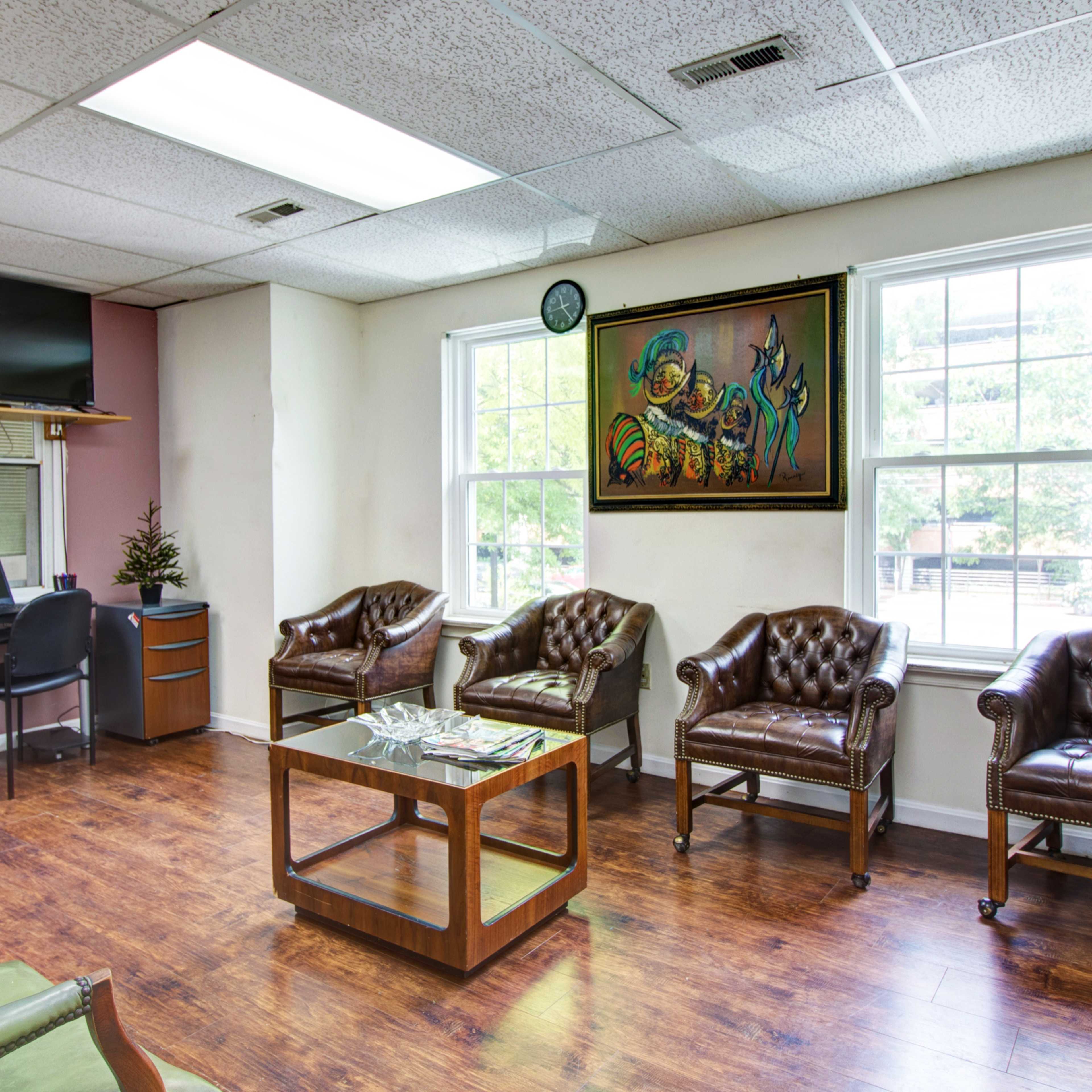 A waiting room with brown leather chairs, a small desk with a monitor, and a colorful painting on the wall.