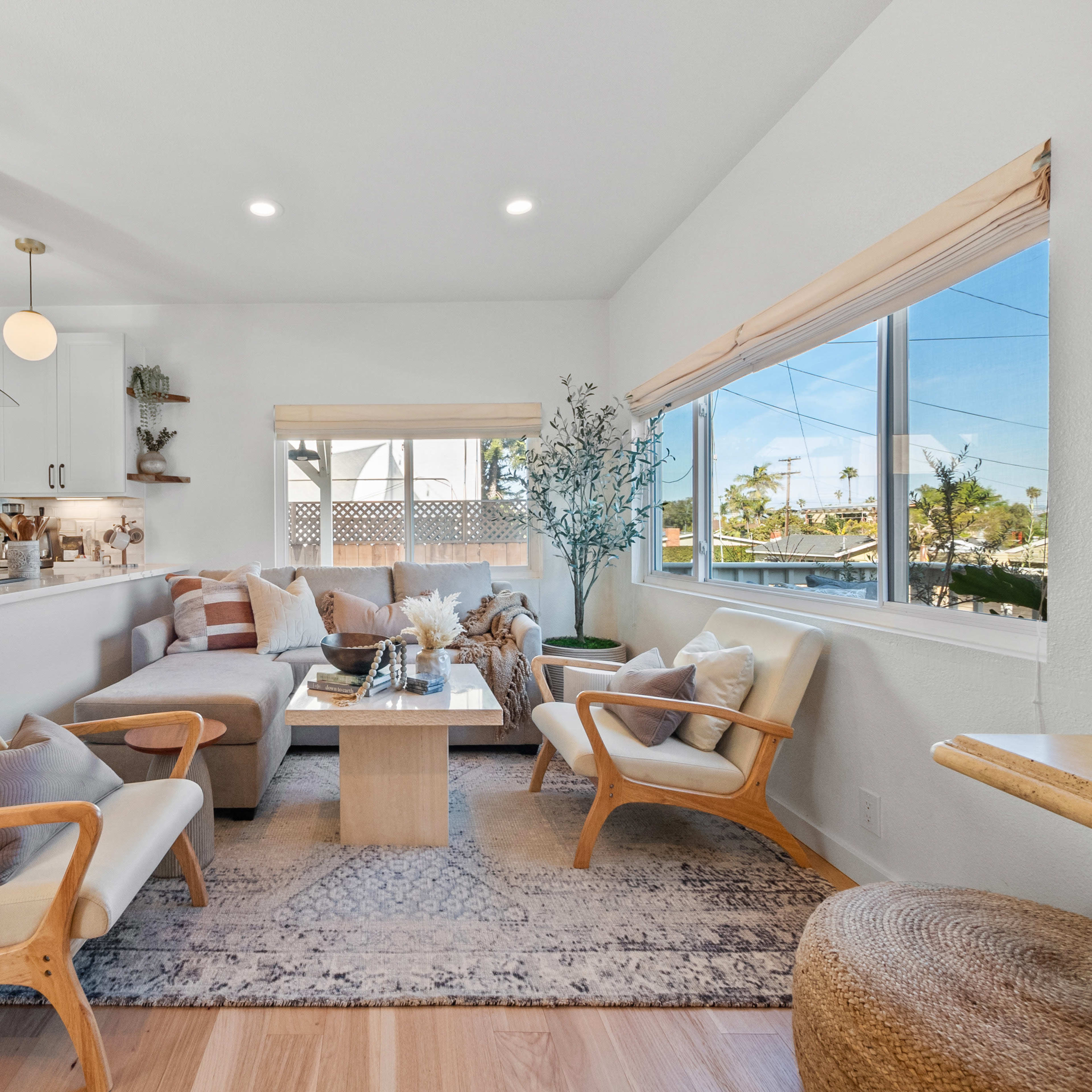 A modern living room features a light-colored sectional sofa, wooden chairs, a coffee table, and large windows with views outside.