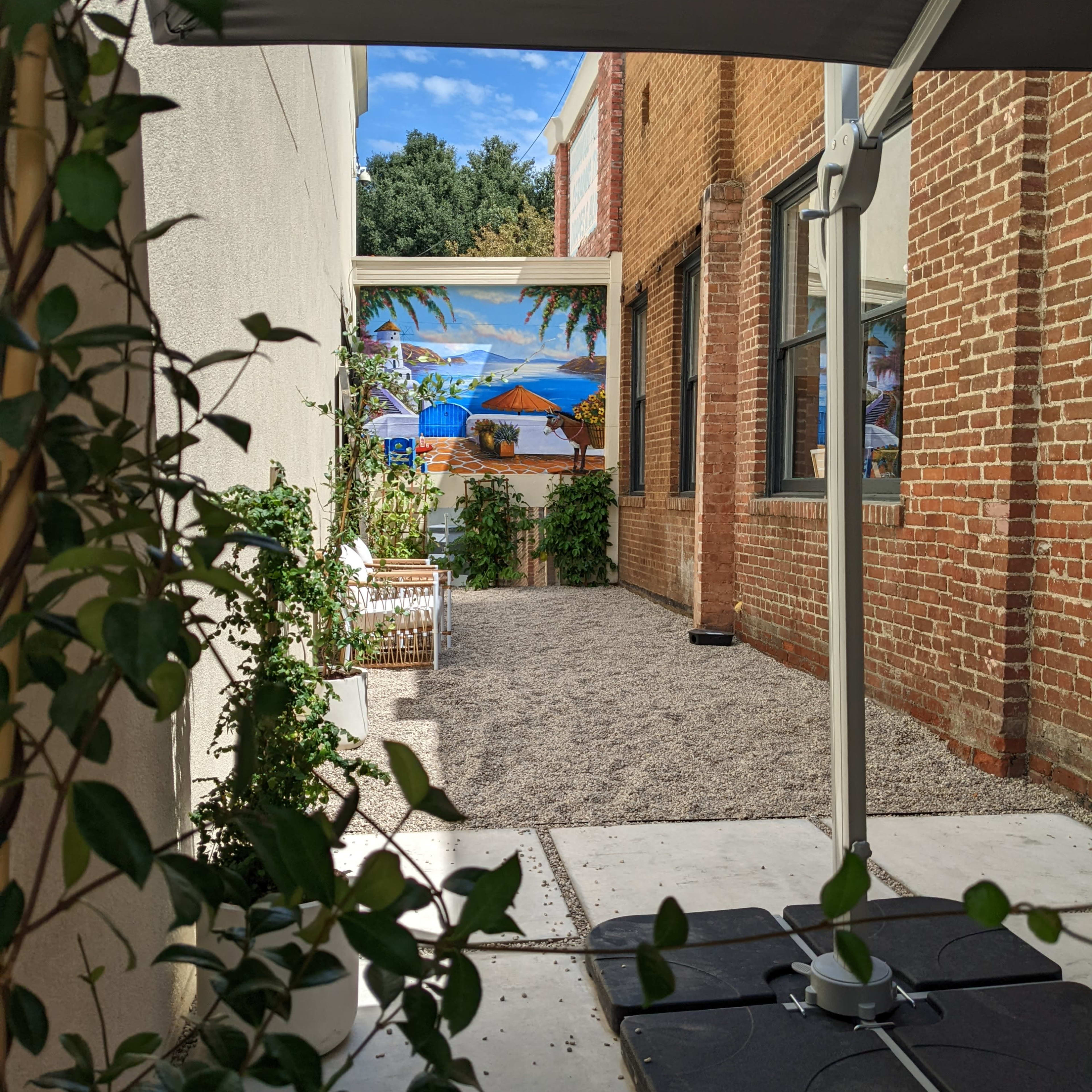 A narrow outdoor space between two brick buildings, featuring gravel ground, a basketball hoop, and a mural of a tropical scene on the wall.