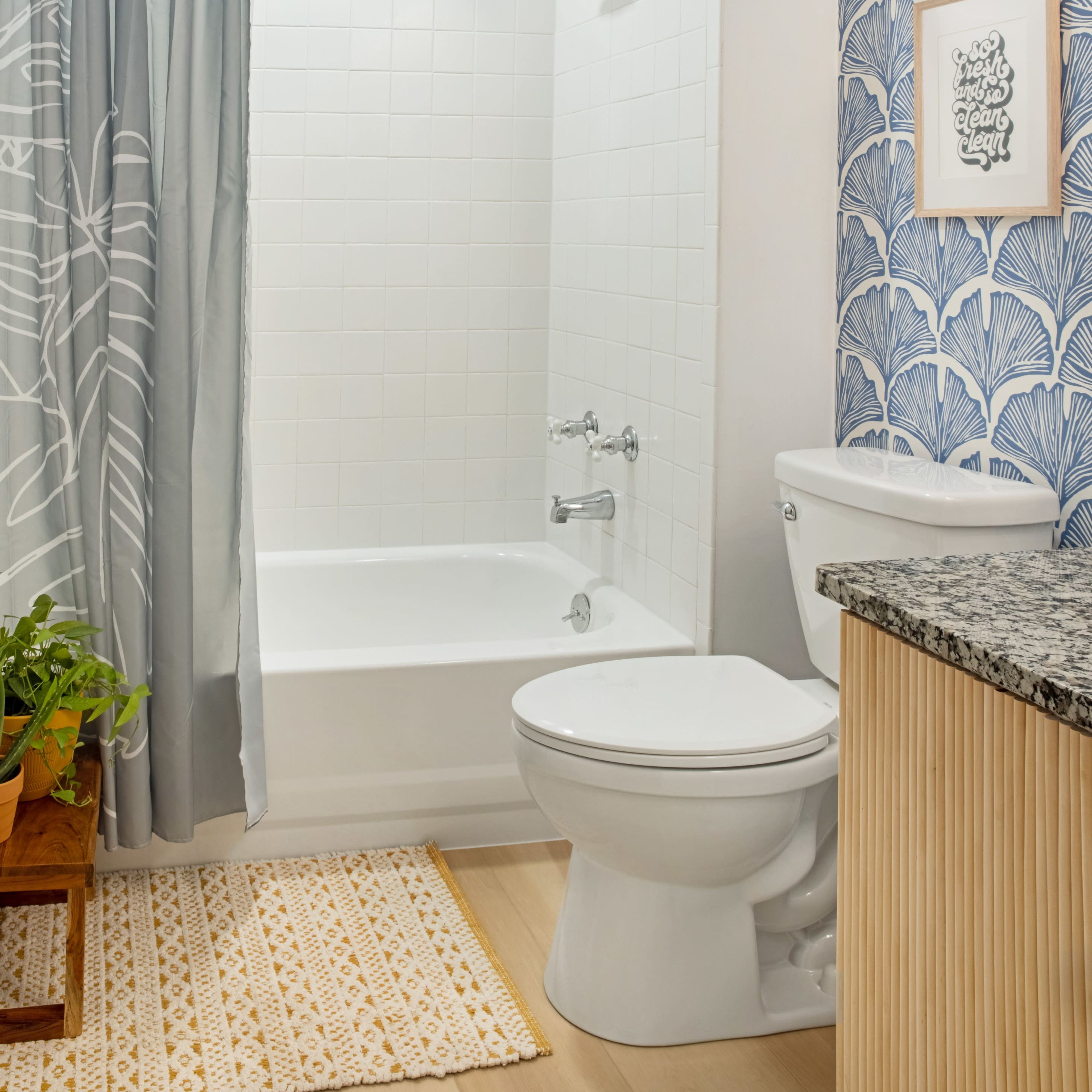 A bathroom featuring a shower-tub combo, a toilet, a granite countertop, and decorative wallpaper with a leaf pattern.