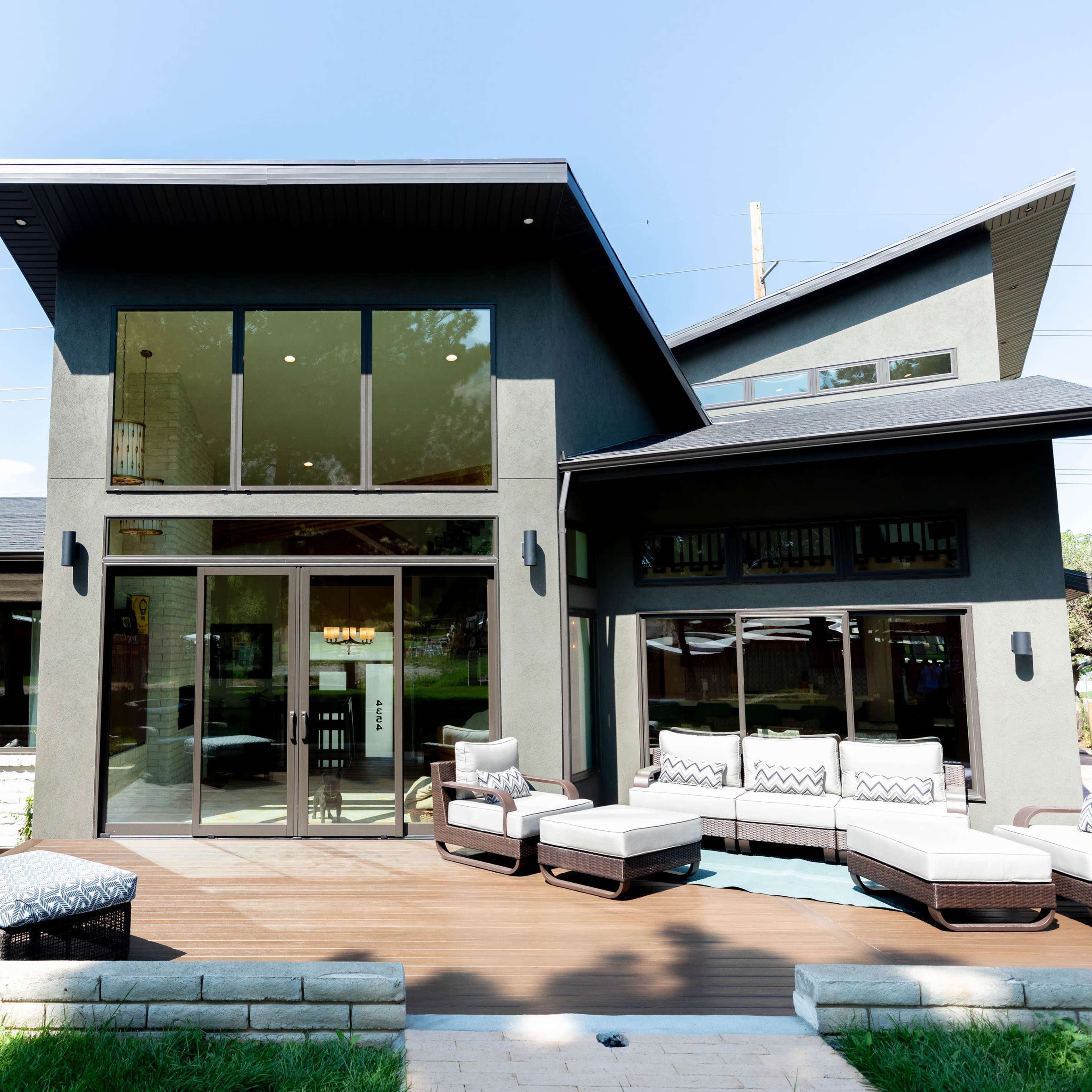 A modern two-story house with large glass windows and a deck furnished with outdoor seating.