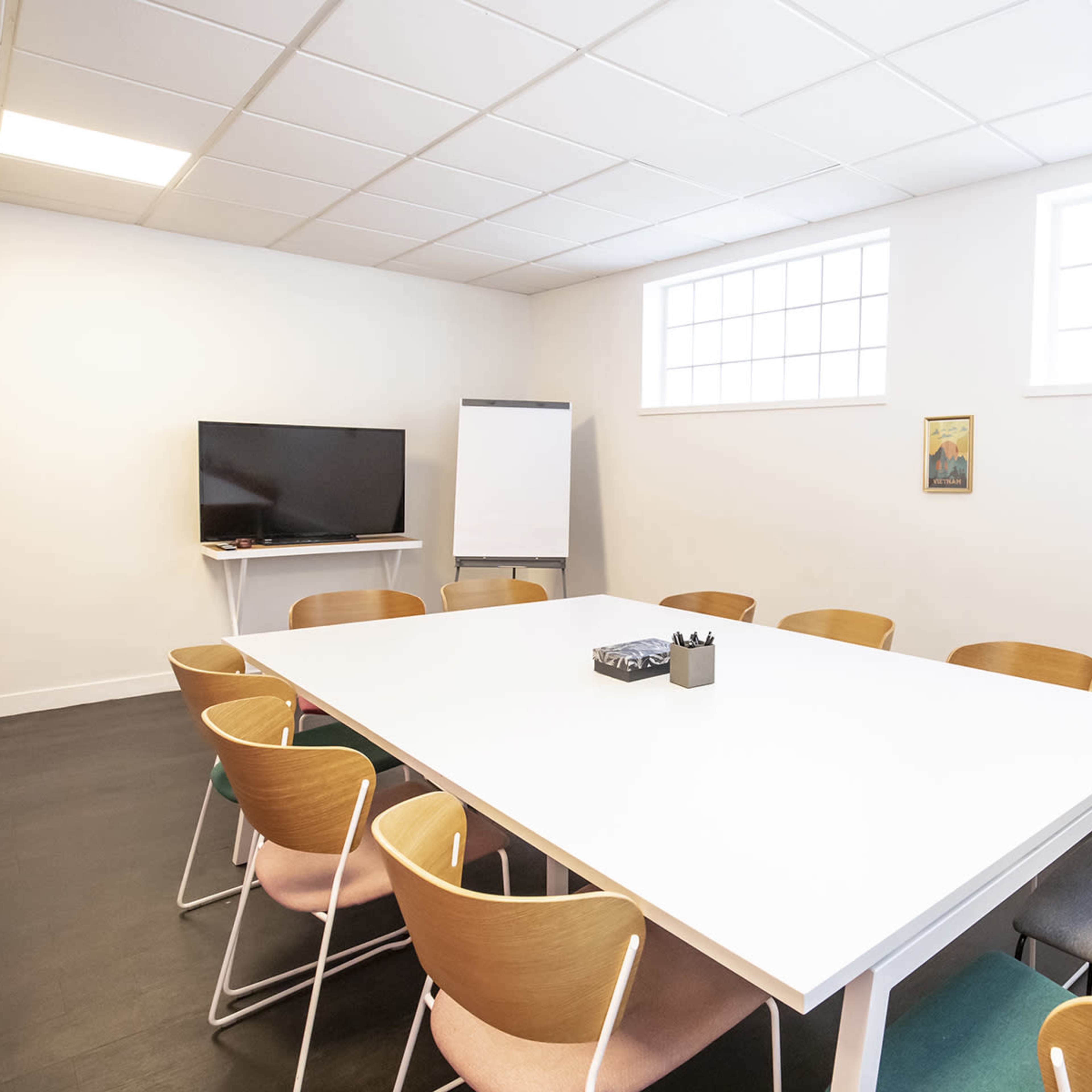 A modern conference room features a large white table surrounded by wooden chairs, with a television and a whiteboard in the corner.
