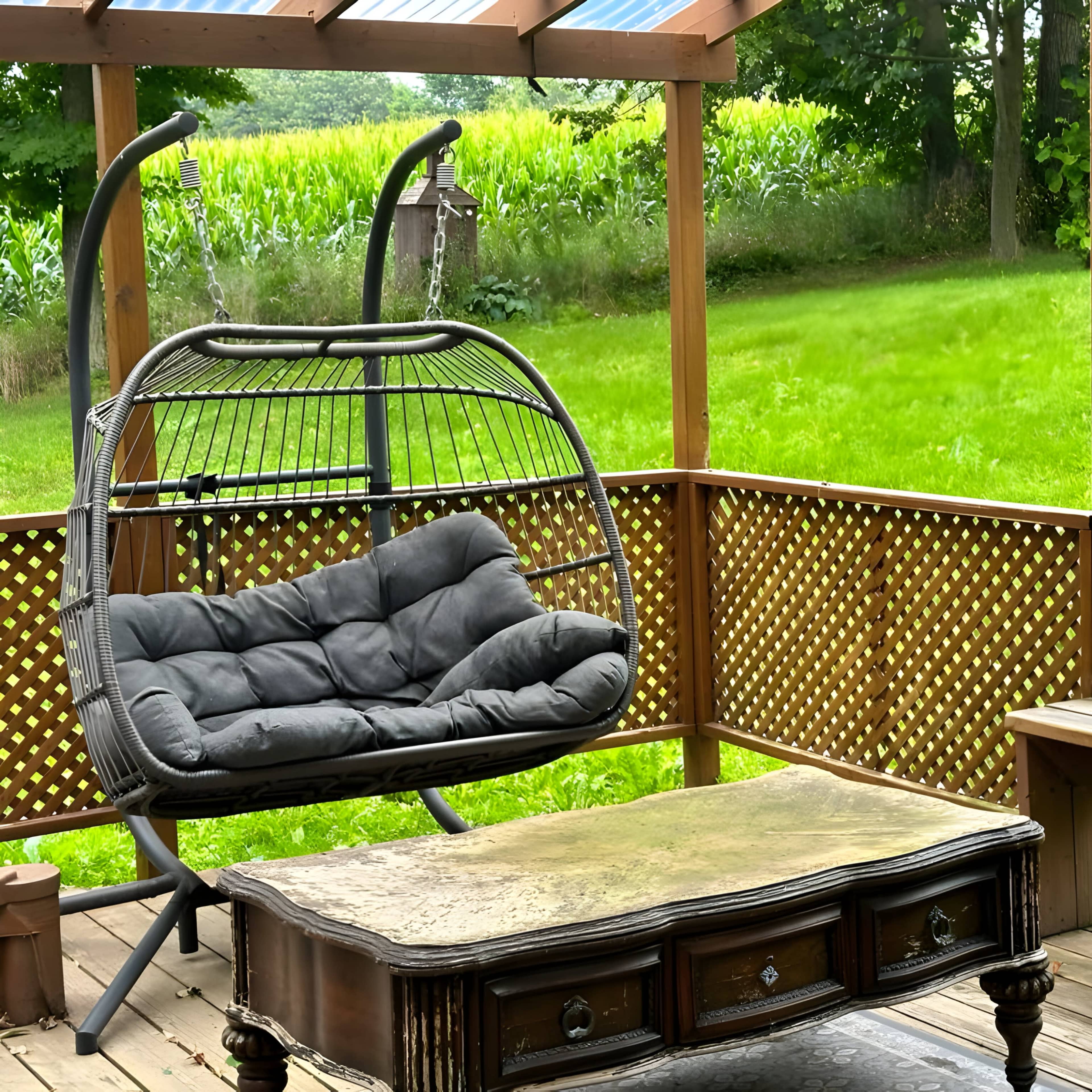 A suspended chair and a wooden coffee table are arranged on a deck surrounded by green grass and trees.