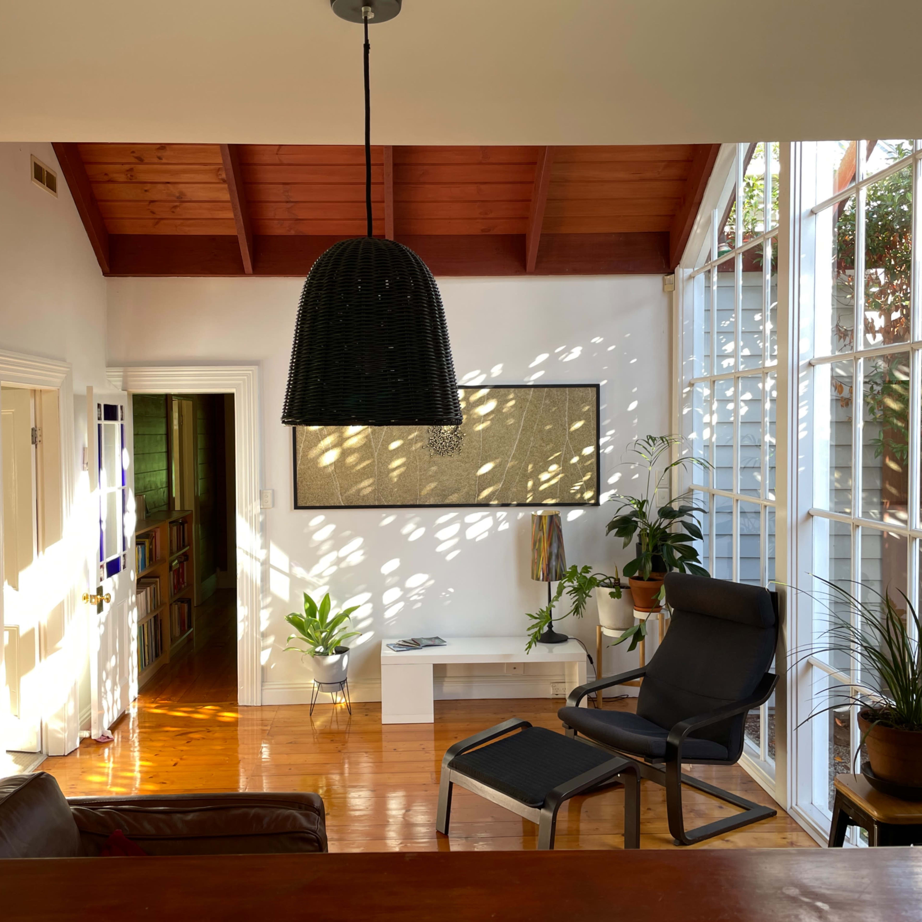 A sunlit room features a black pendant light, a lounge chair, potted plants, and large windows, with wooden ceiling beams overhead.