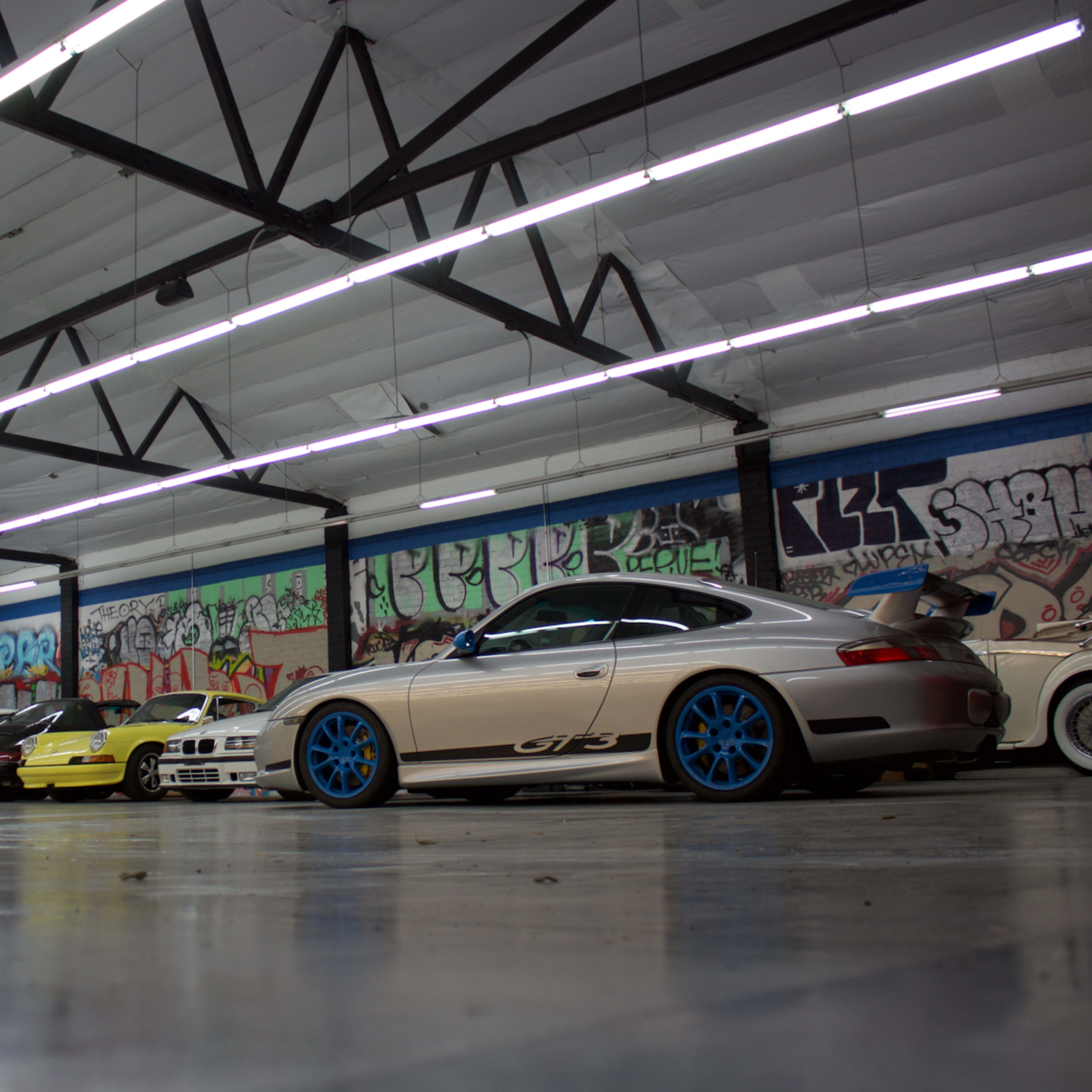 A row of various cars, including a Porsche with blue wheels, is parked in an industrial space adorned with colorful graffiti.