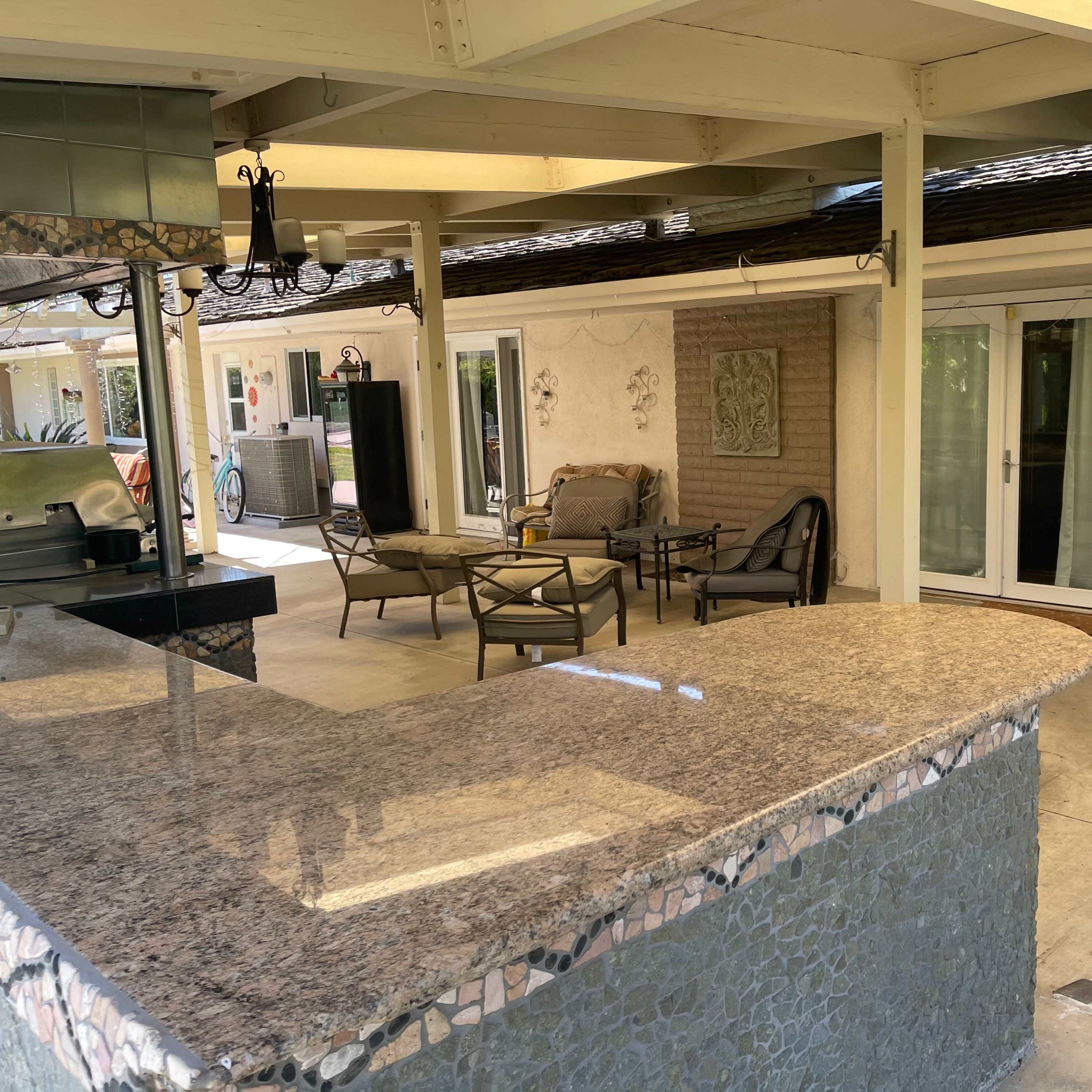 A covered outdoor kitchen with a granite countertop and seating area adjacent to a house.