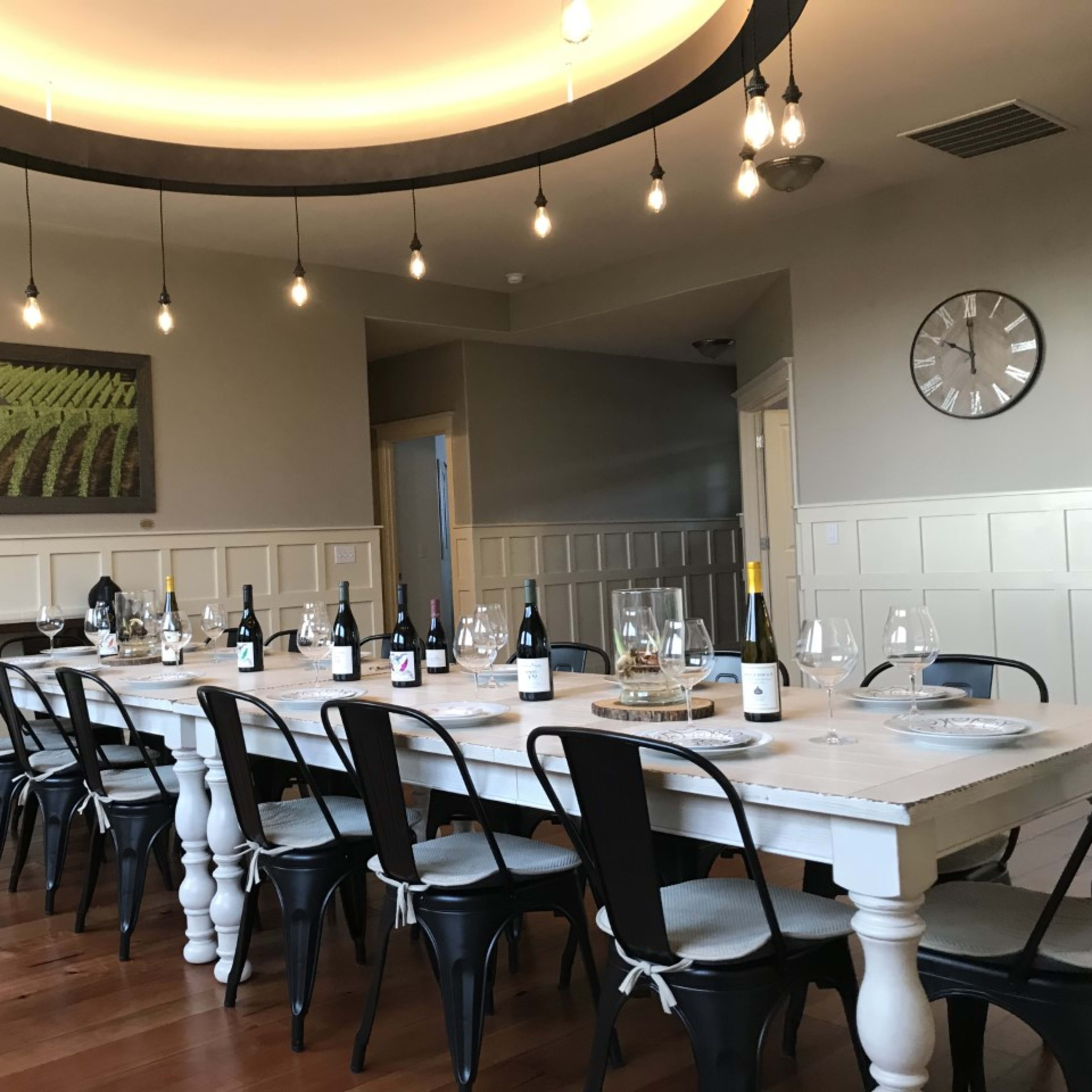 A long, white dining table with wine bottles and glasses is set in a well-lit room featuring a vineyard painting on the wall.