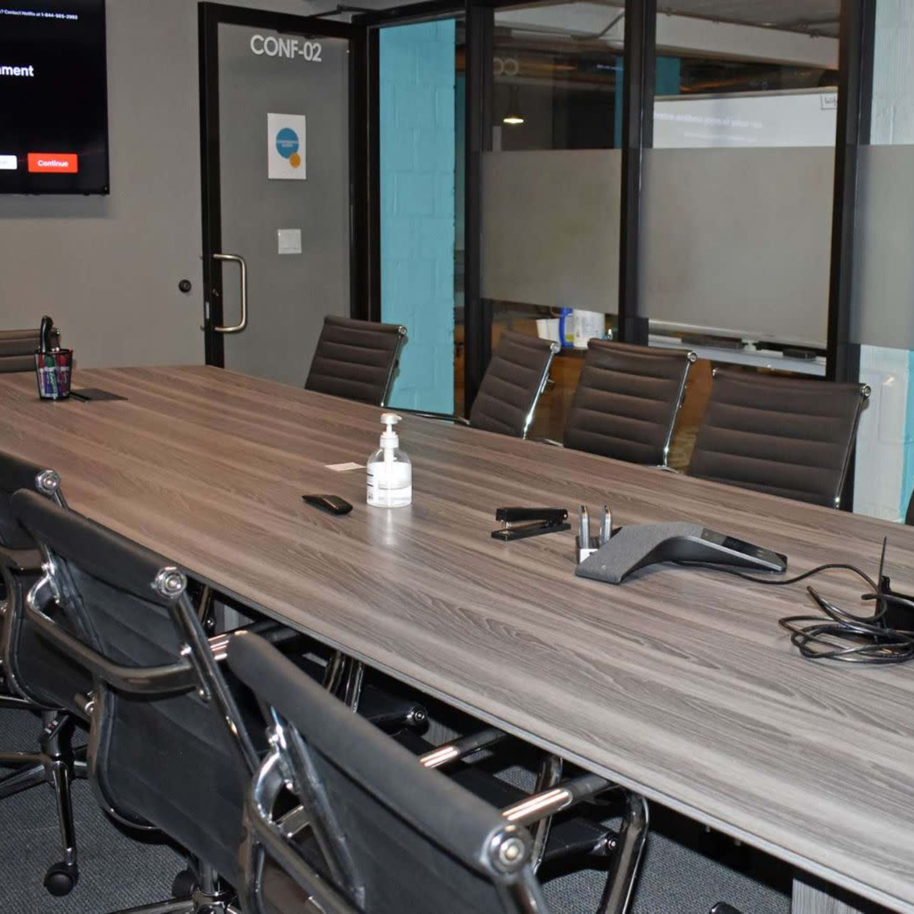 A large conference room features a long wooden table surrounded by black rolling chairs, equipped with a phone and various office supplies, and a screen displaying information on the wall.