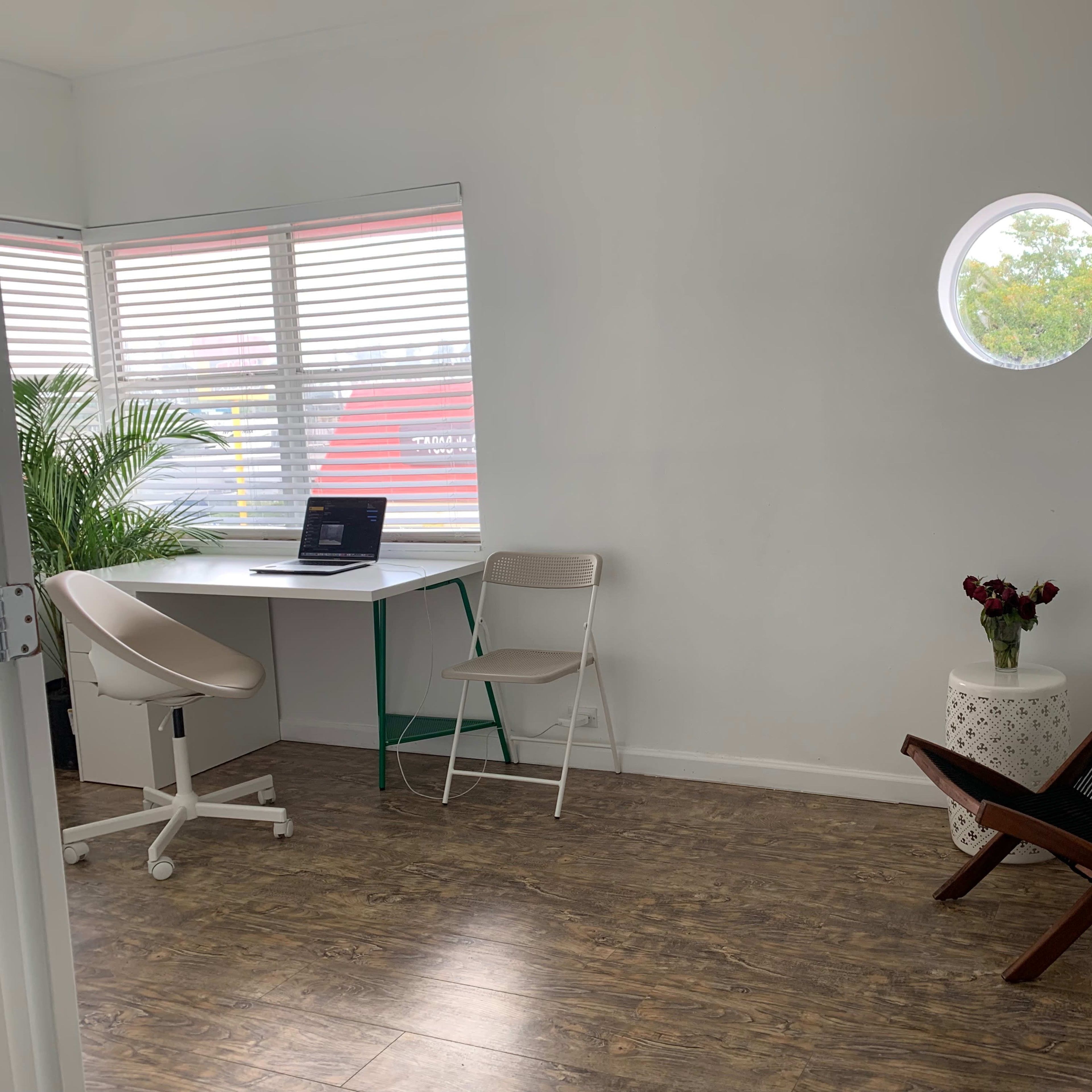 A bright, minimalistic room with a round window, a small desk with a laptop, a chair, a plant, and a decorative table with flowers.