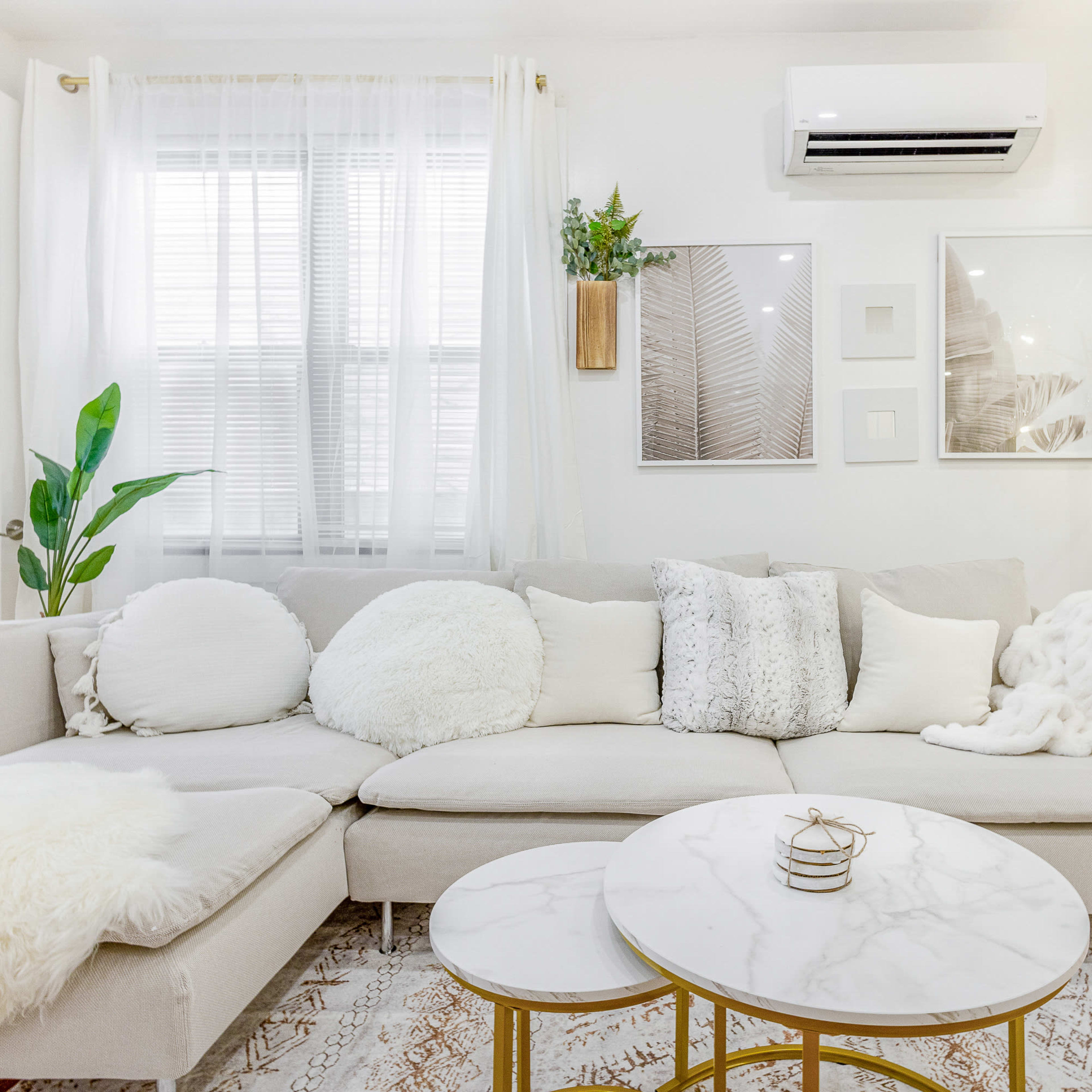 A bright living room with a light-colored sectional sofa, decorative pillows, and a marble-topped coffee table set against a backdrop of white walls and large windows.