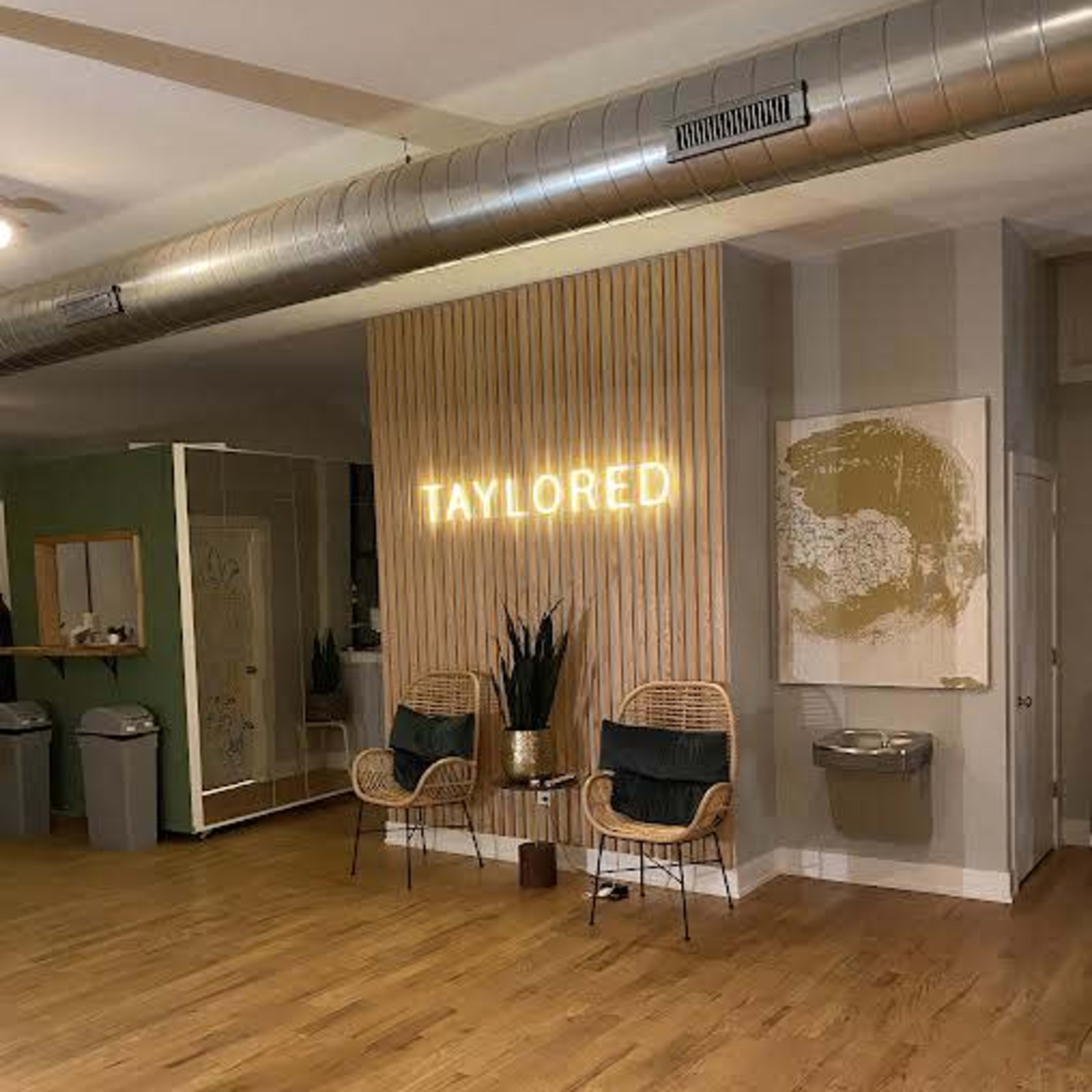 A spacious room with wooden floors, featuring a neon sign that reads "TAYLORED," two rattan chairs, and a small table, along with a water fountain and decorative plants.