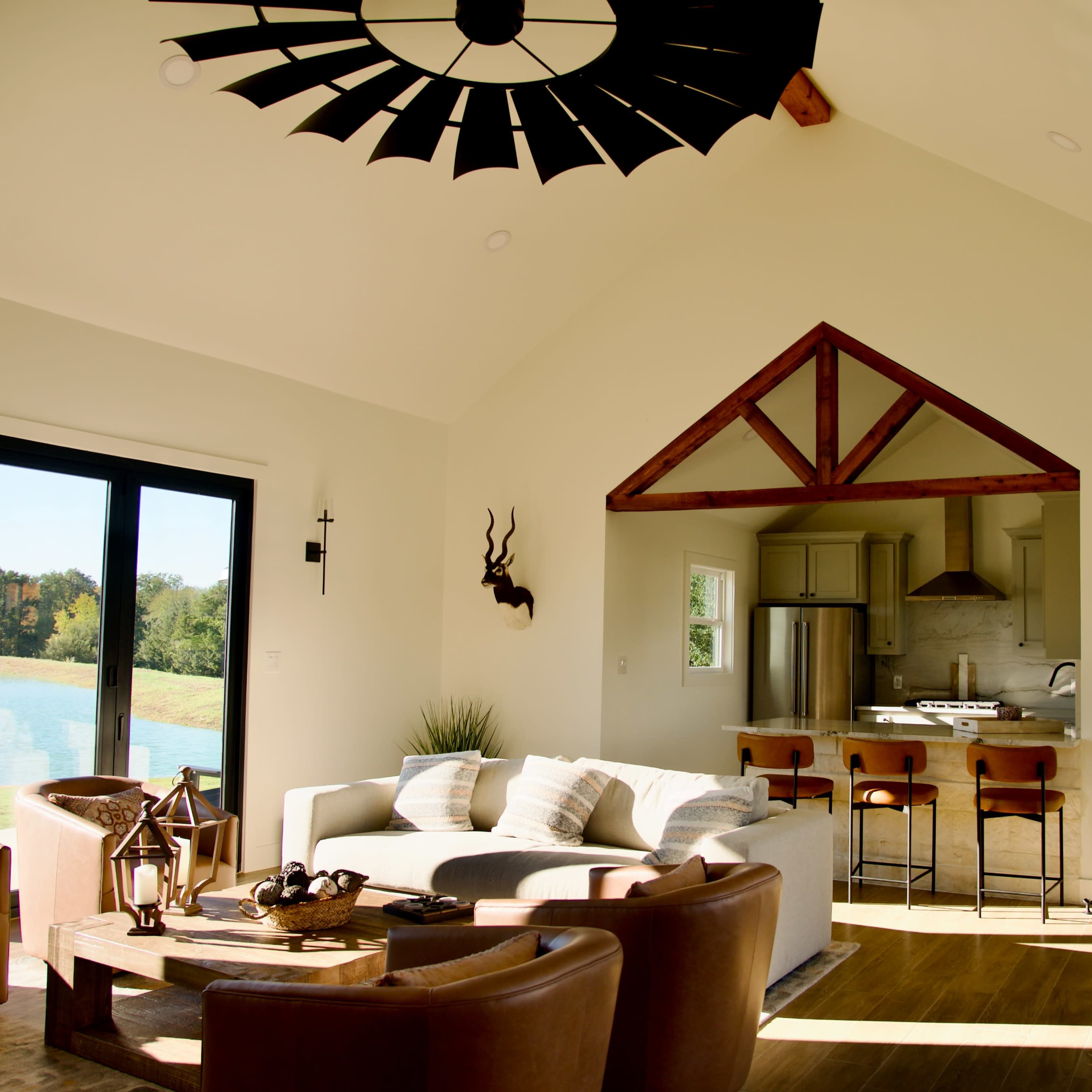 A spacious, modern living room features a tall ceiling, large windows overlooking a lake, and a kitchen area with wooden accents.