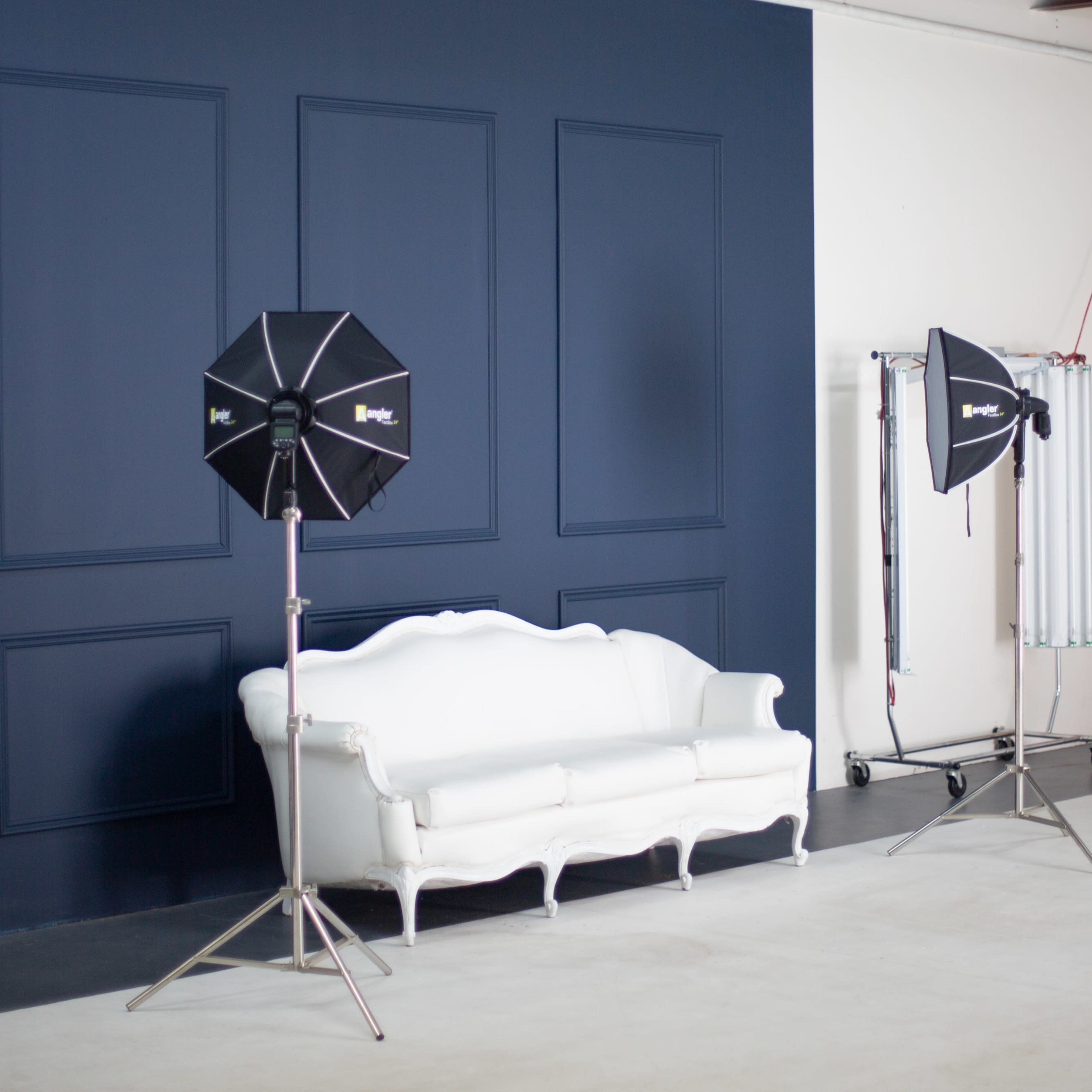 A photography studio with a white couch, two light reflectors on stands, and a dark blue wall.