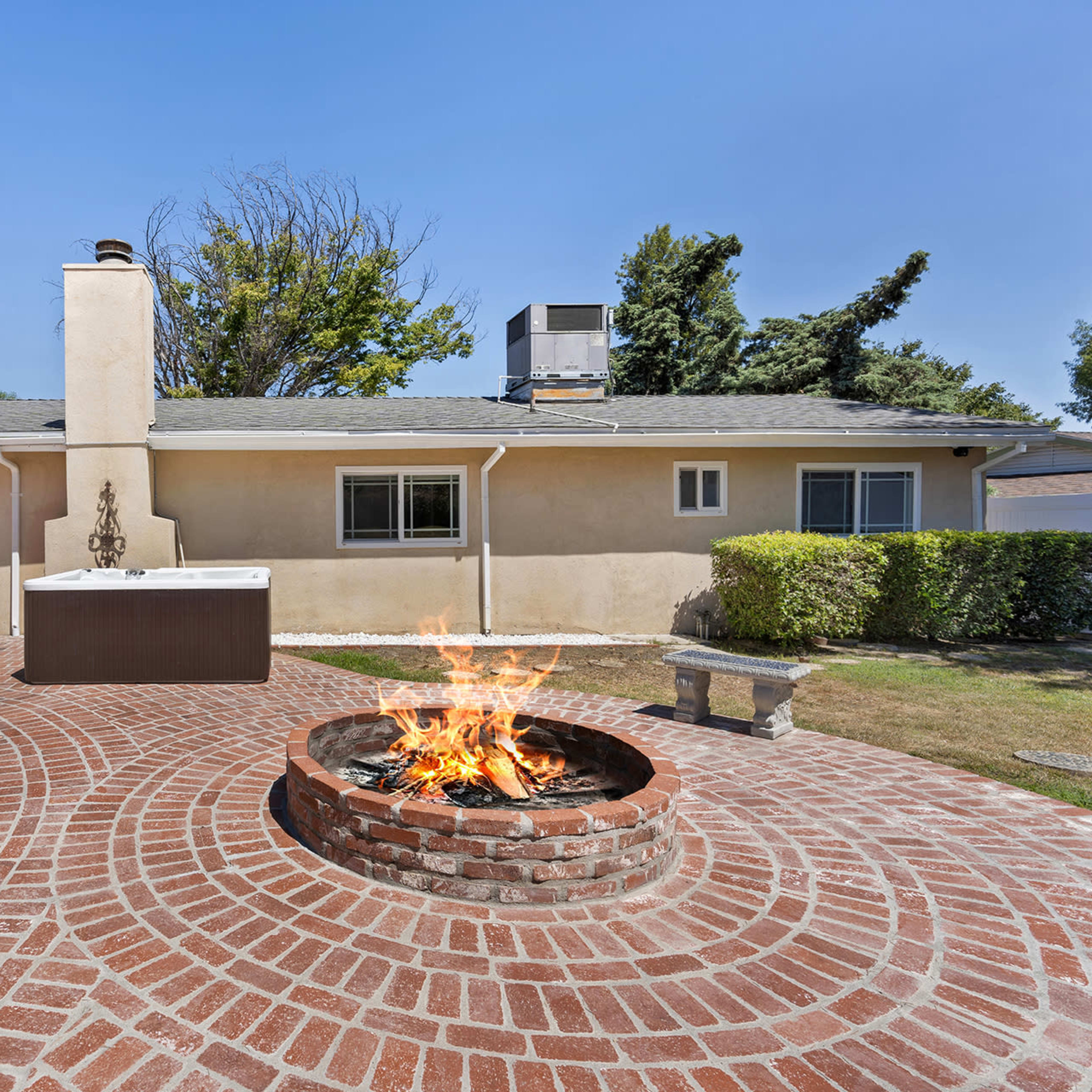 A brick patio with a fire pit is situated in front of a single-story house surrounded by greenery.