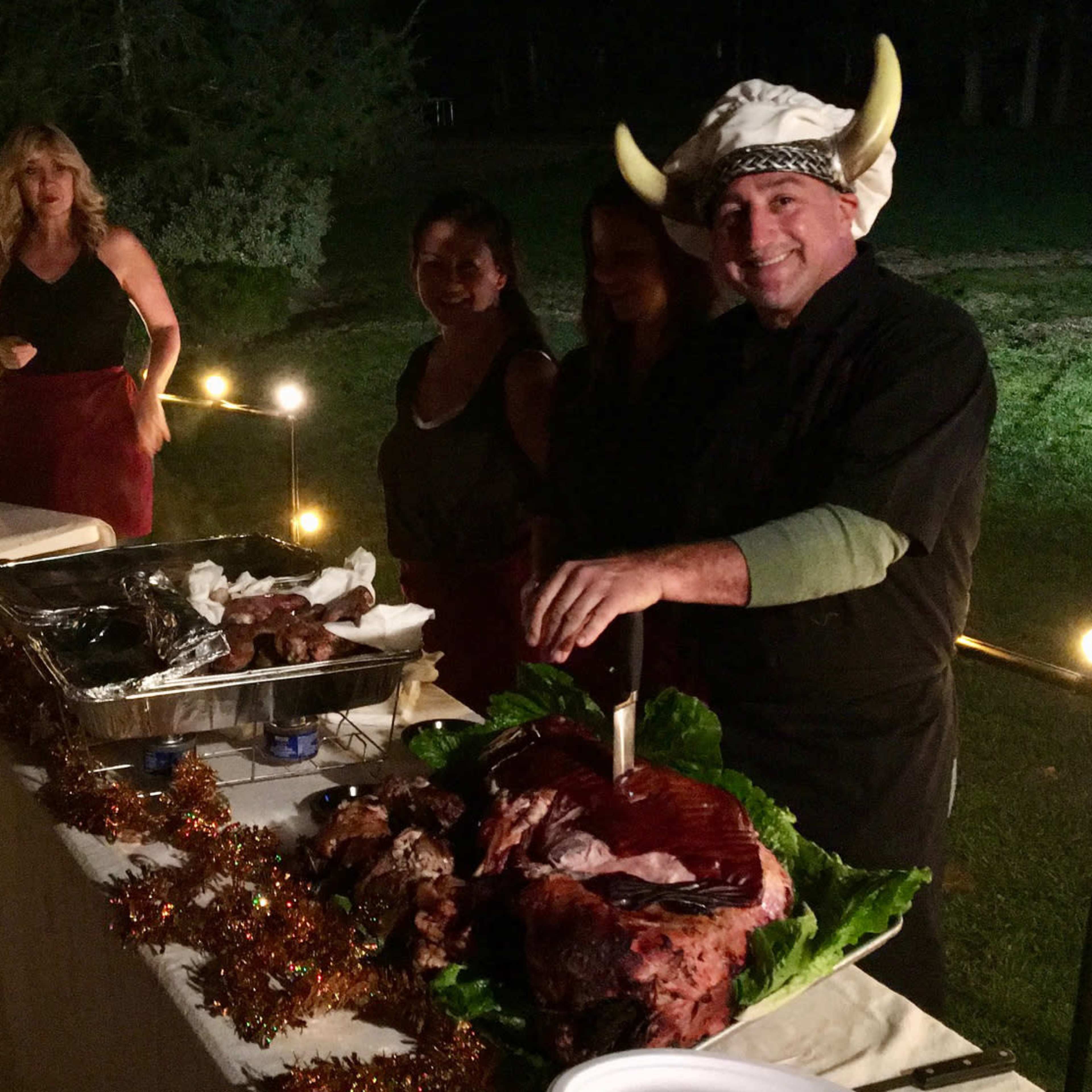 A man wearing a chef's hat and viking horns carves a large roast on a table, while several women stand nearby in an outdoor setting with lights strung above.