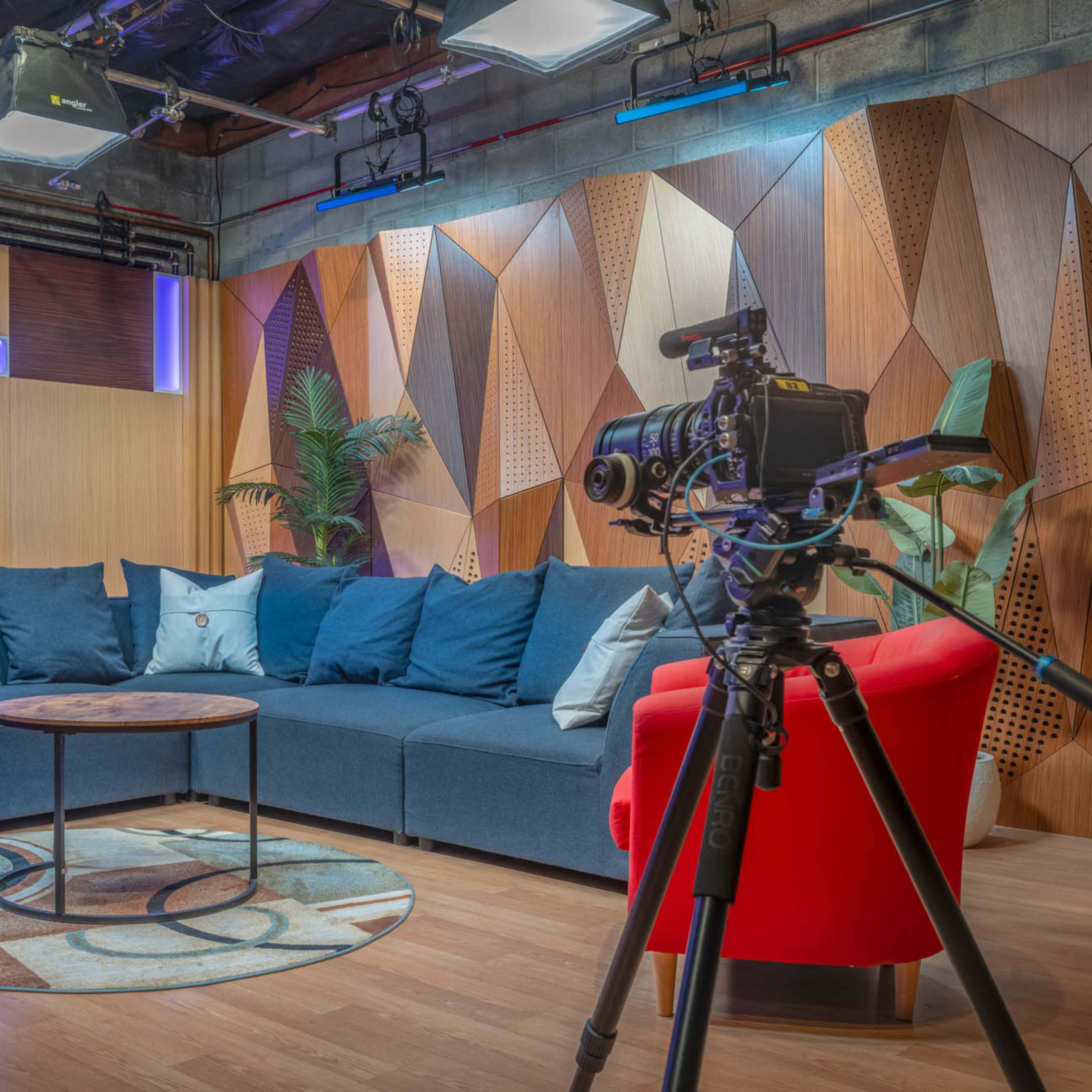 A modern studio set with a blue sofa, a round coffee table, and a camera on a tripod, surrounded by geometric wooden panels and accent lighting.