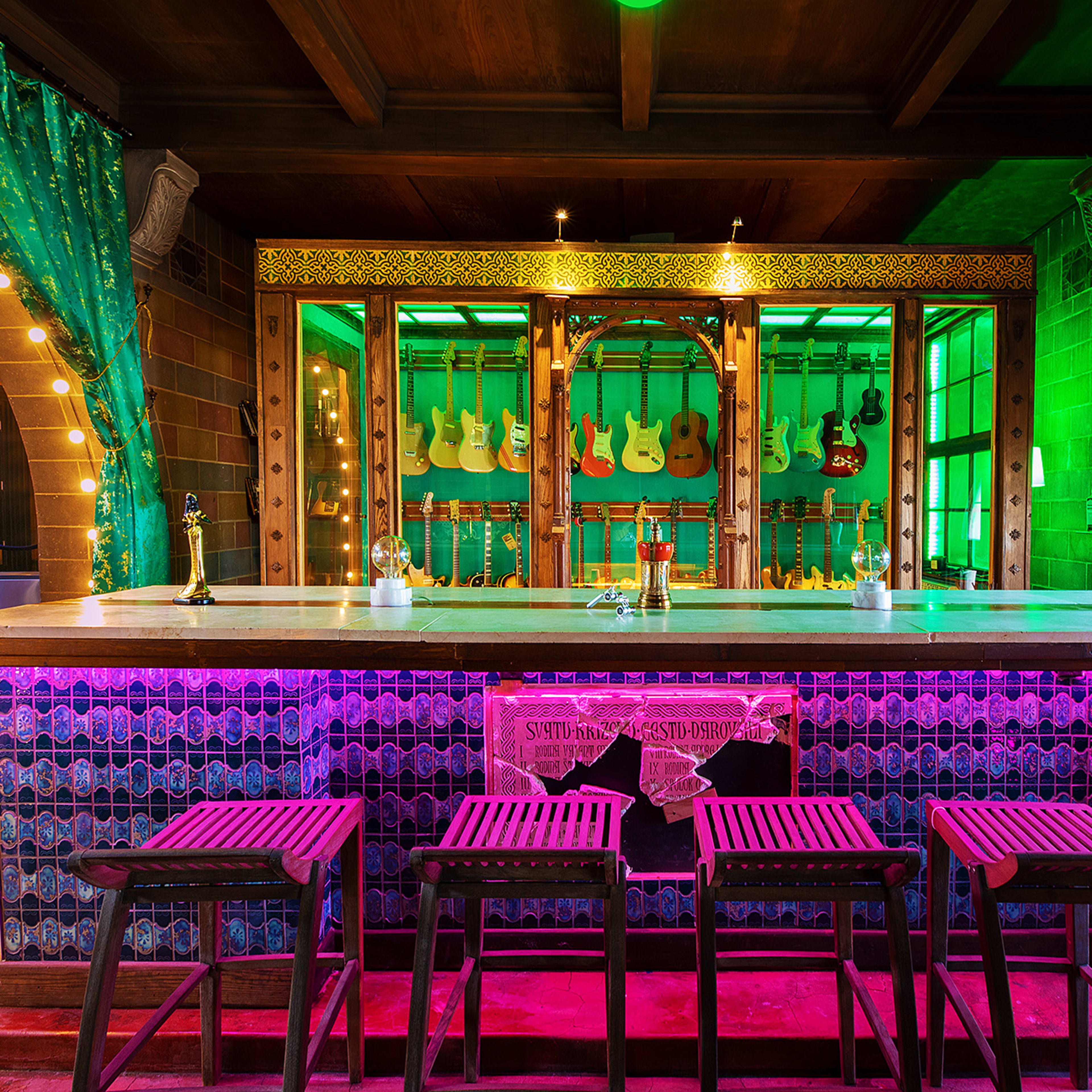A colorful bar area featuring a wooden counter, several bar stools, and a display of guitars in a vibrant setting with green and purple lighting.