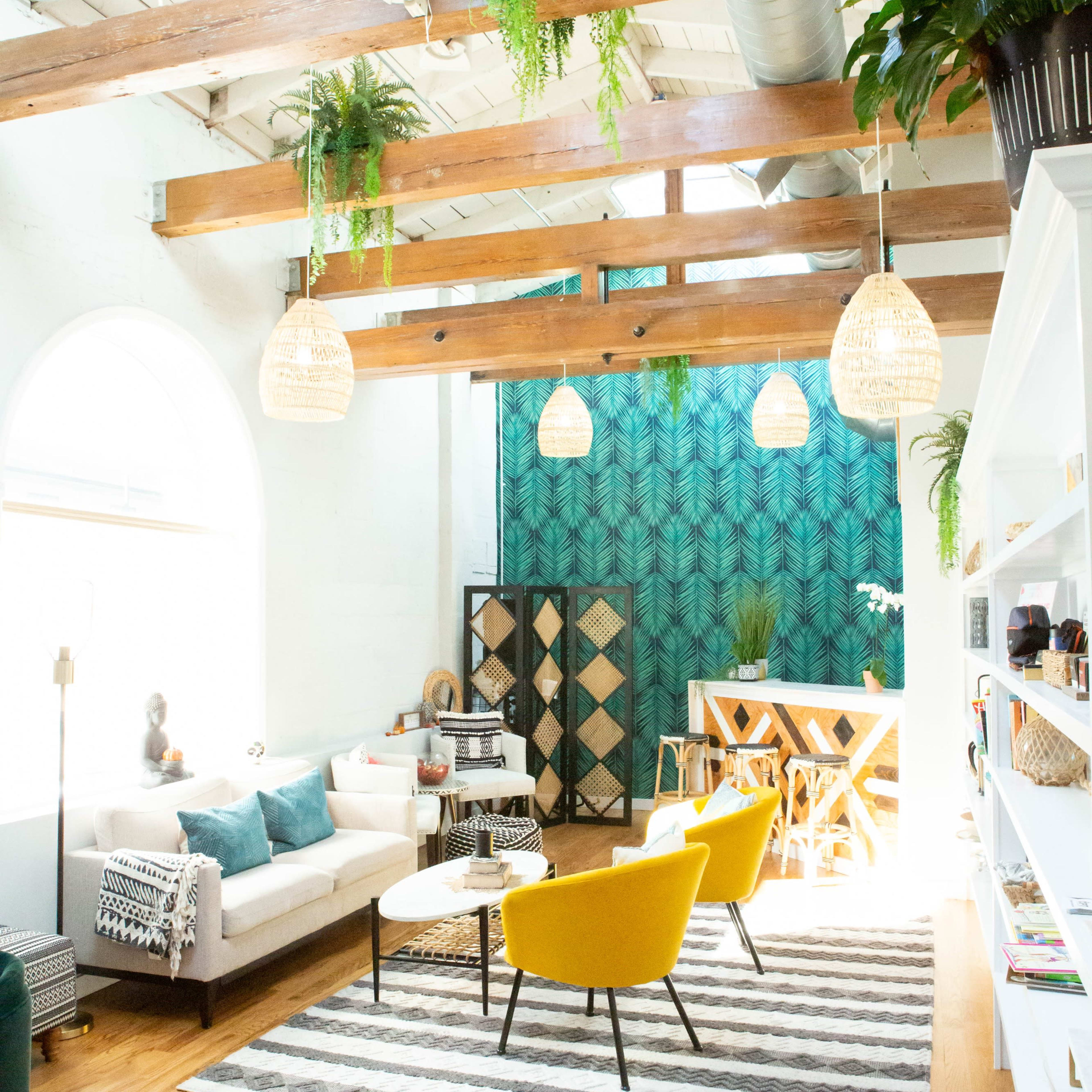The bright and airy living space features a mix of modern furniture, including a white sofa, yellow chairs, and a patterned area rug, with large windows and greenery enhancing the atmosphere.