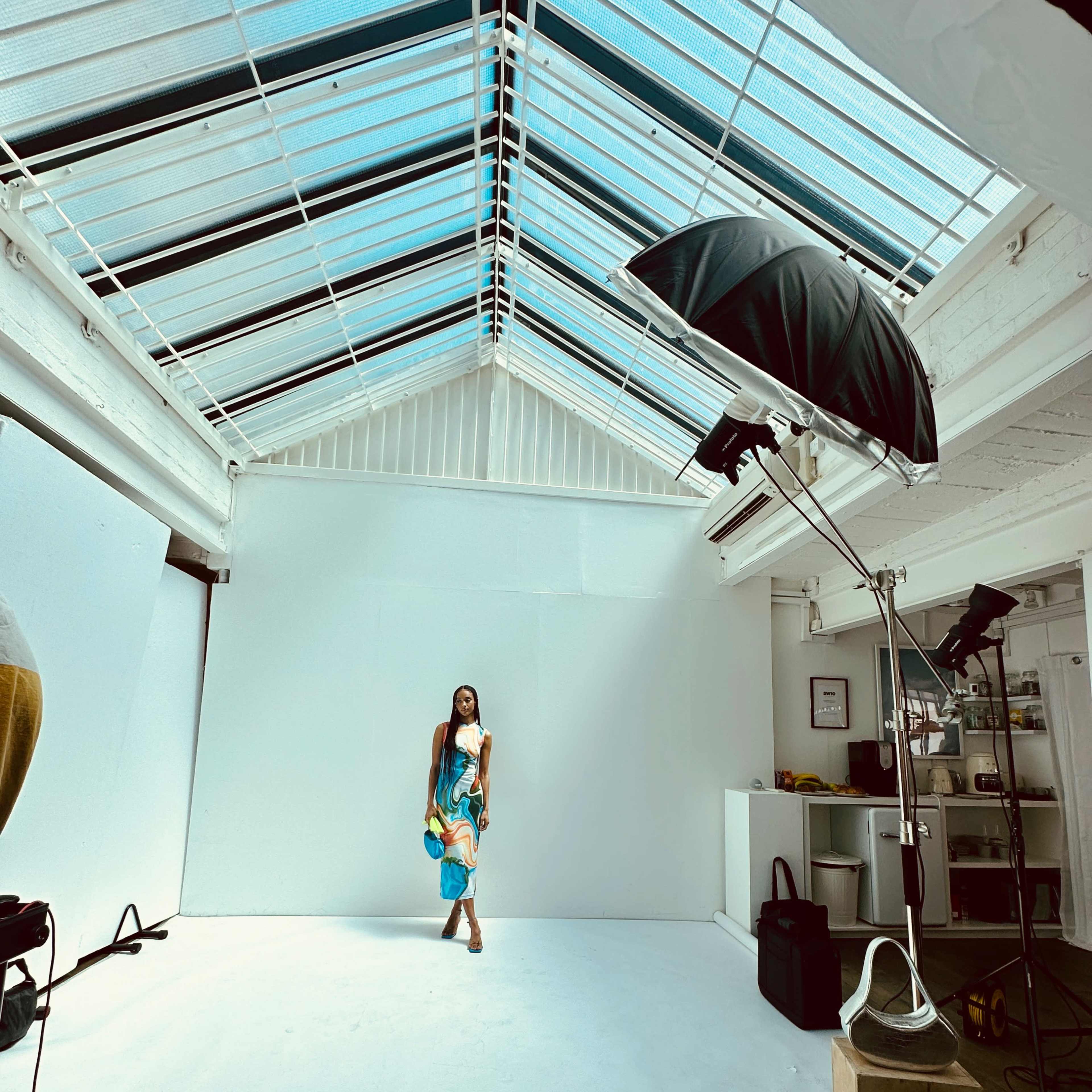 A model holds blue props while standing in a bright, empty photography studio with a slanted glass roof.