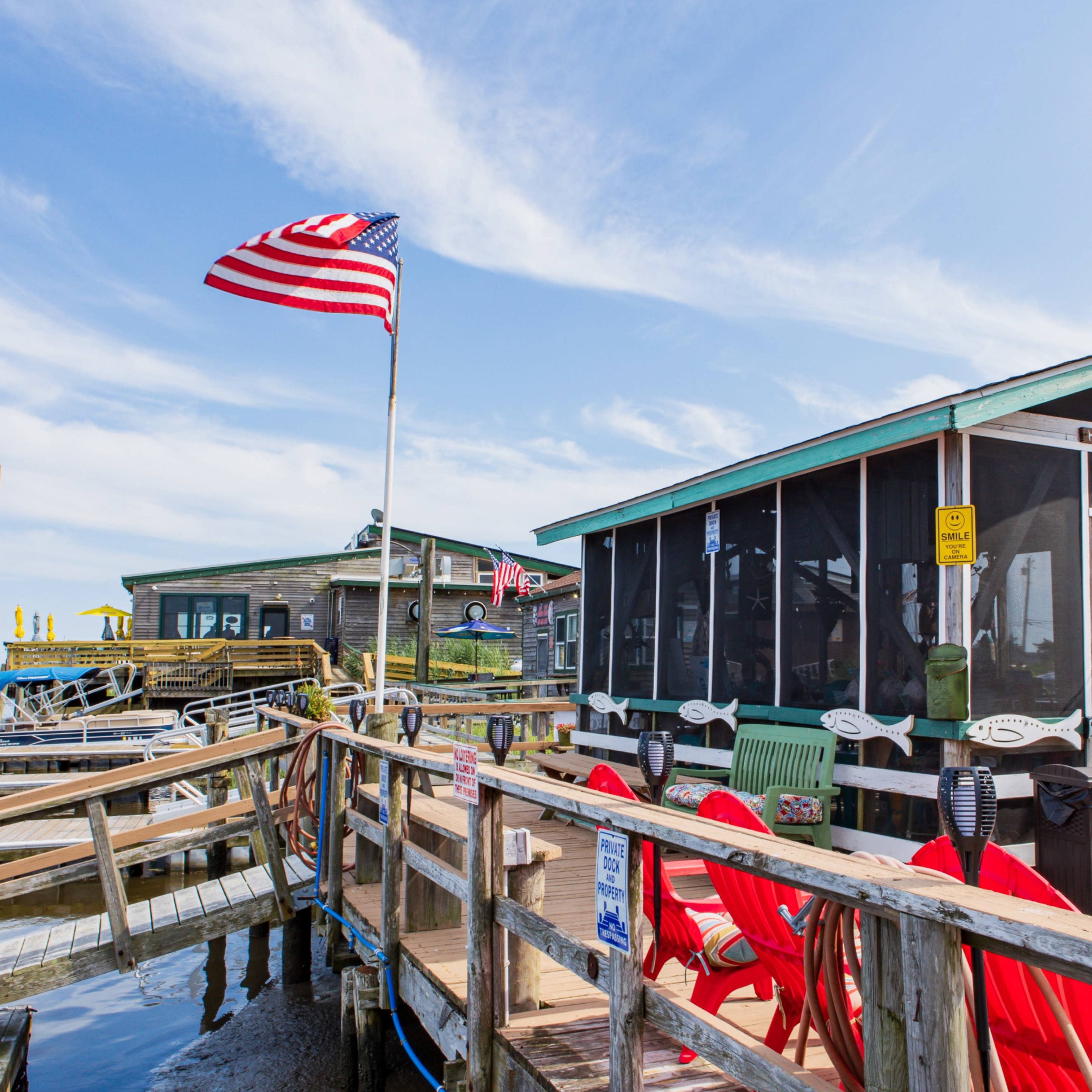 A marina with a wooden dock leading to a screened building, an American flag flying above, and chairs lining the pathway.