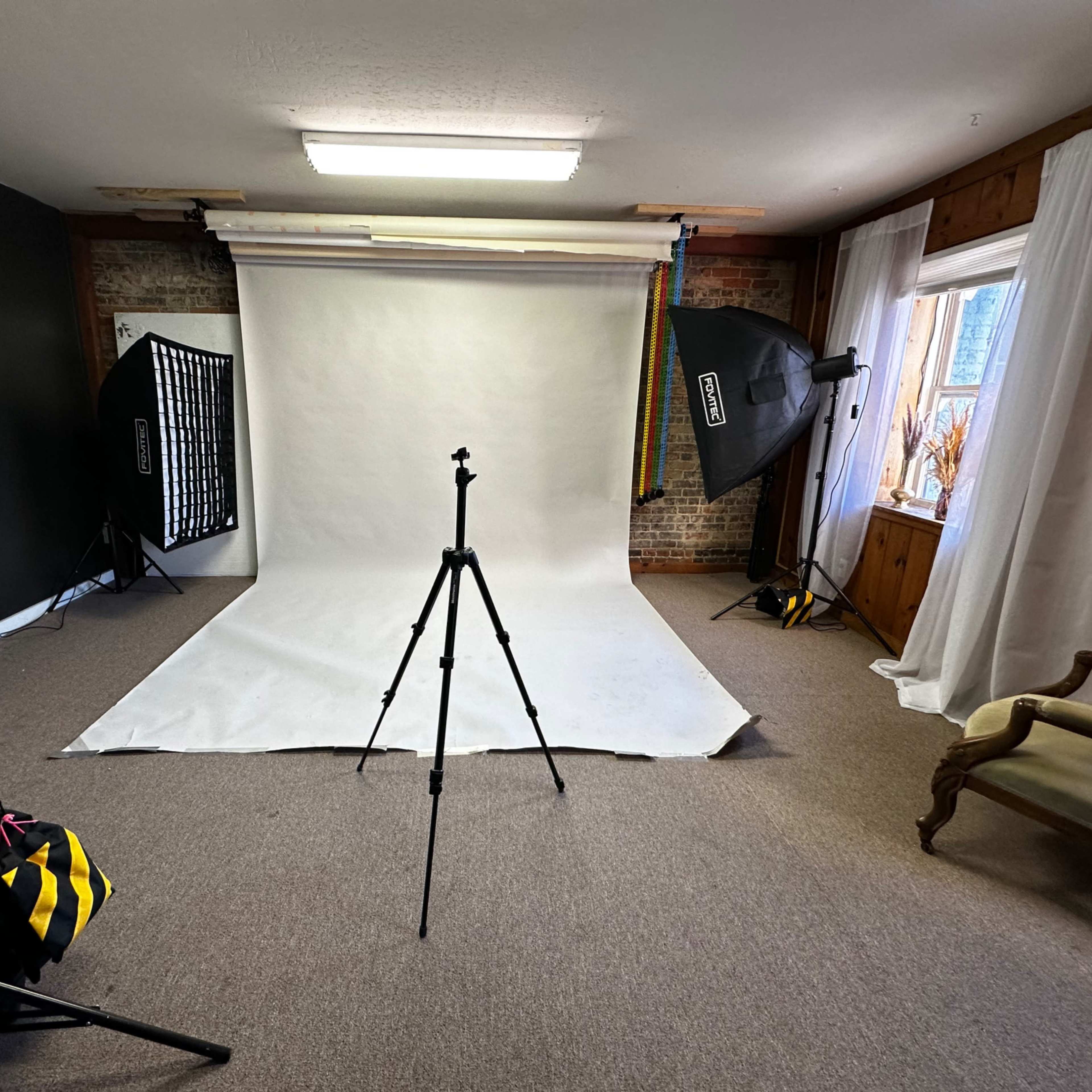 A photography studio setup with a white backdrop, lighting equipment on both sides, and a tripod positioned in the center.