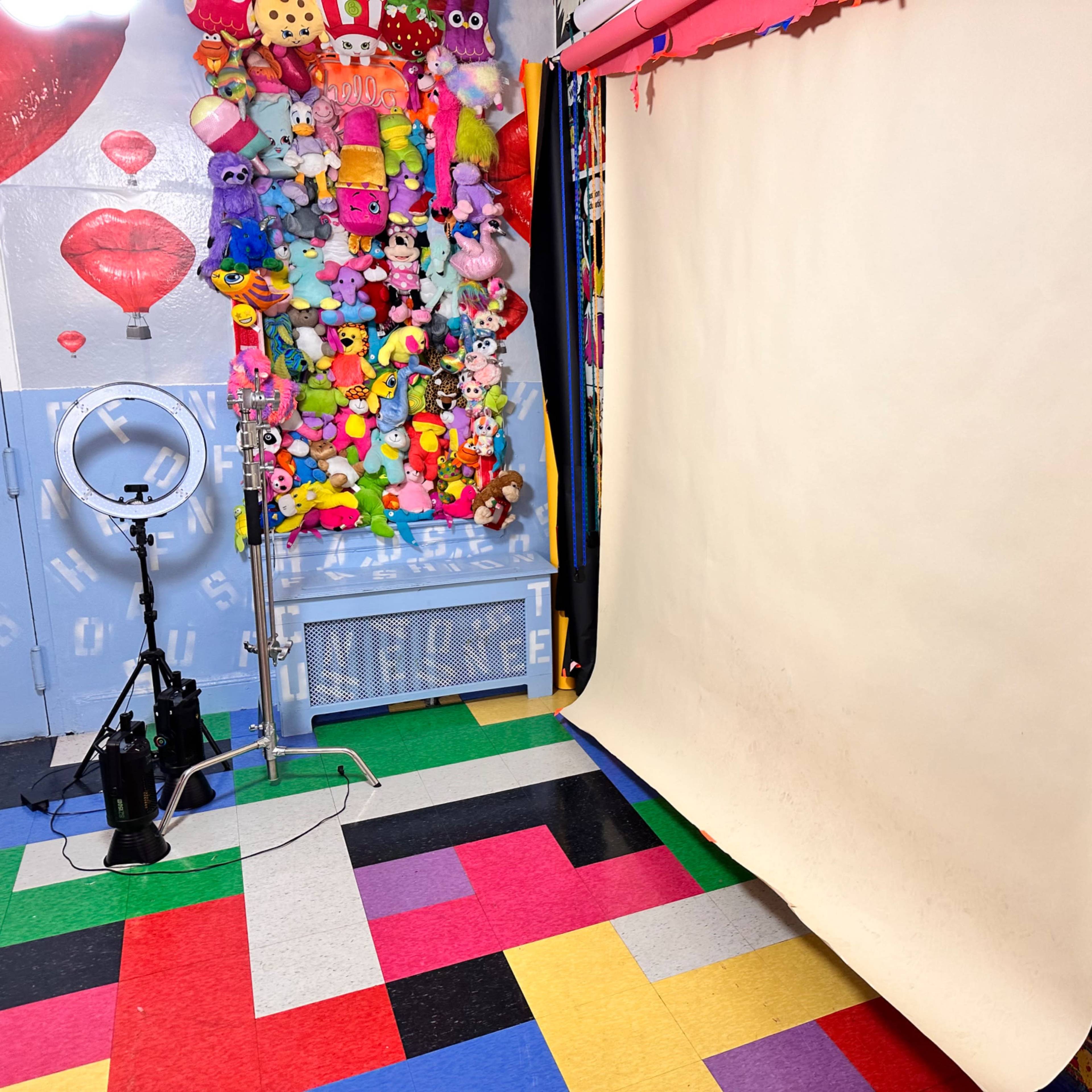 A brightly colored photography studio with a seamless backdrop, a ring light, and walls adorned with colorful decorations and plush toys.