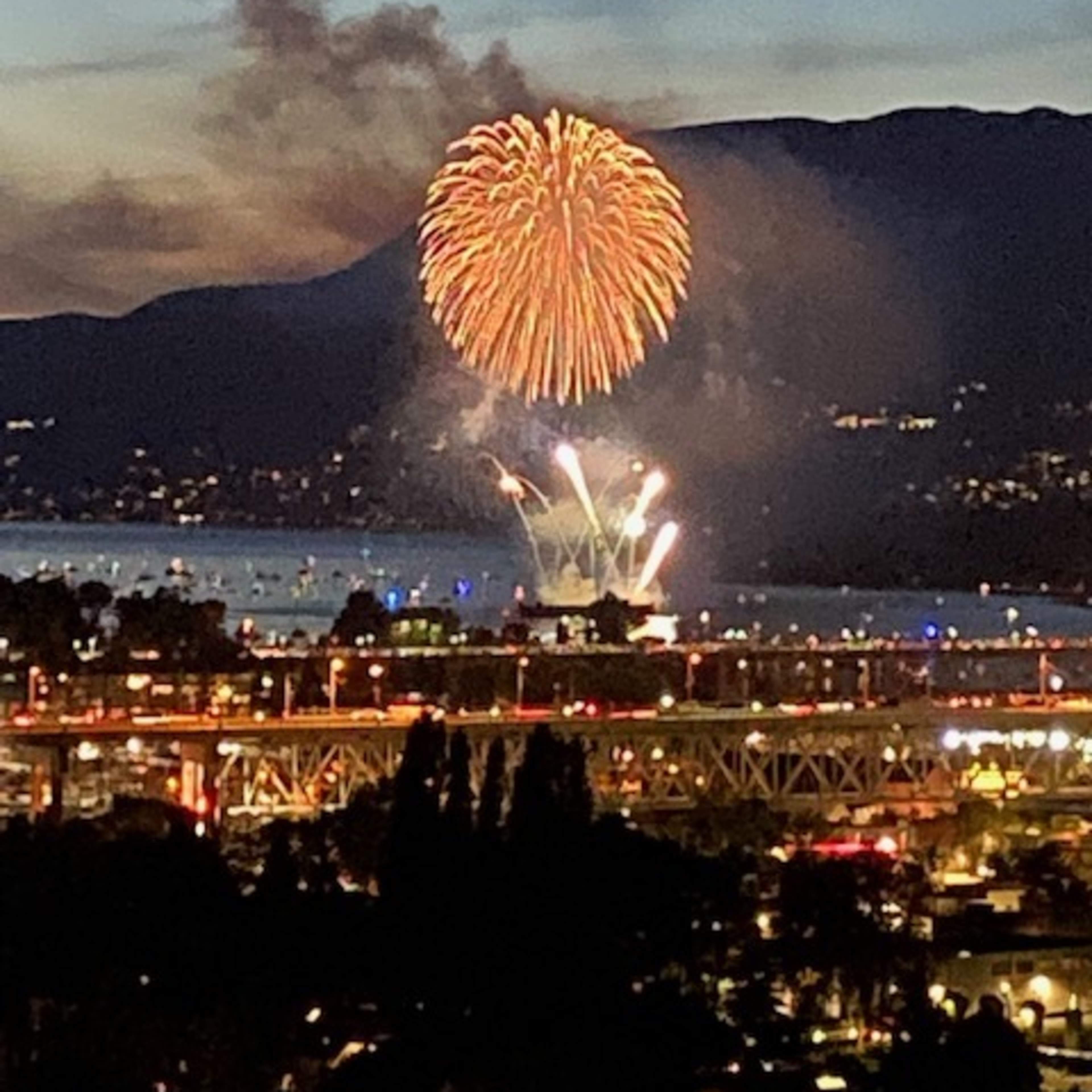 Fireworks illuminate the sky over a body of water, with mountains in the background and a cityscape in the foreground.