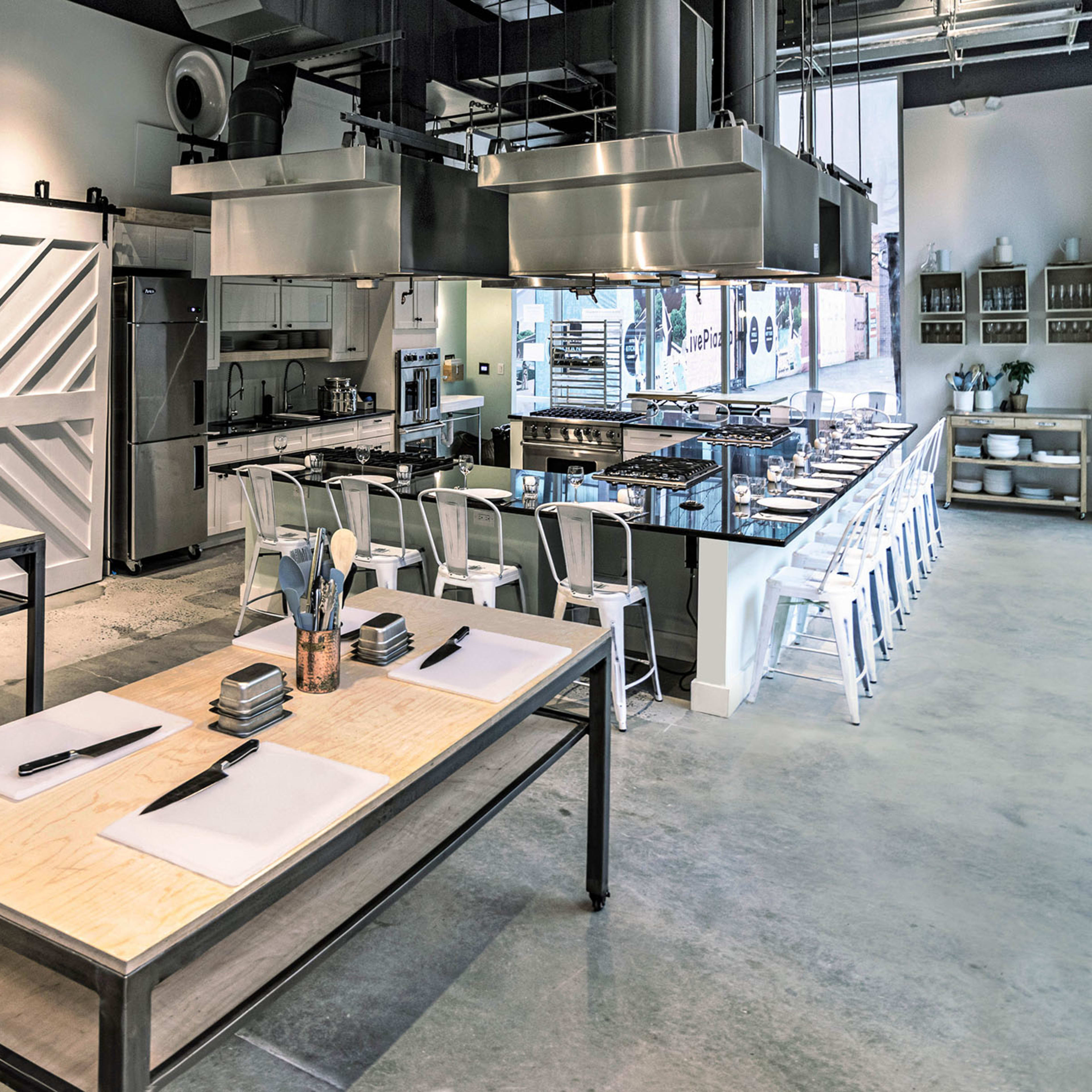 A modern kitchen workspace features metal tables, kitchen equipment, and a row of stools against a minimalist design backdrop.