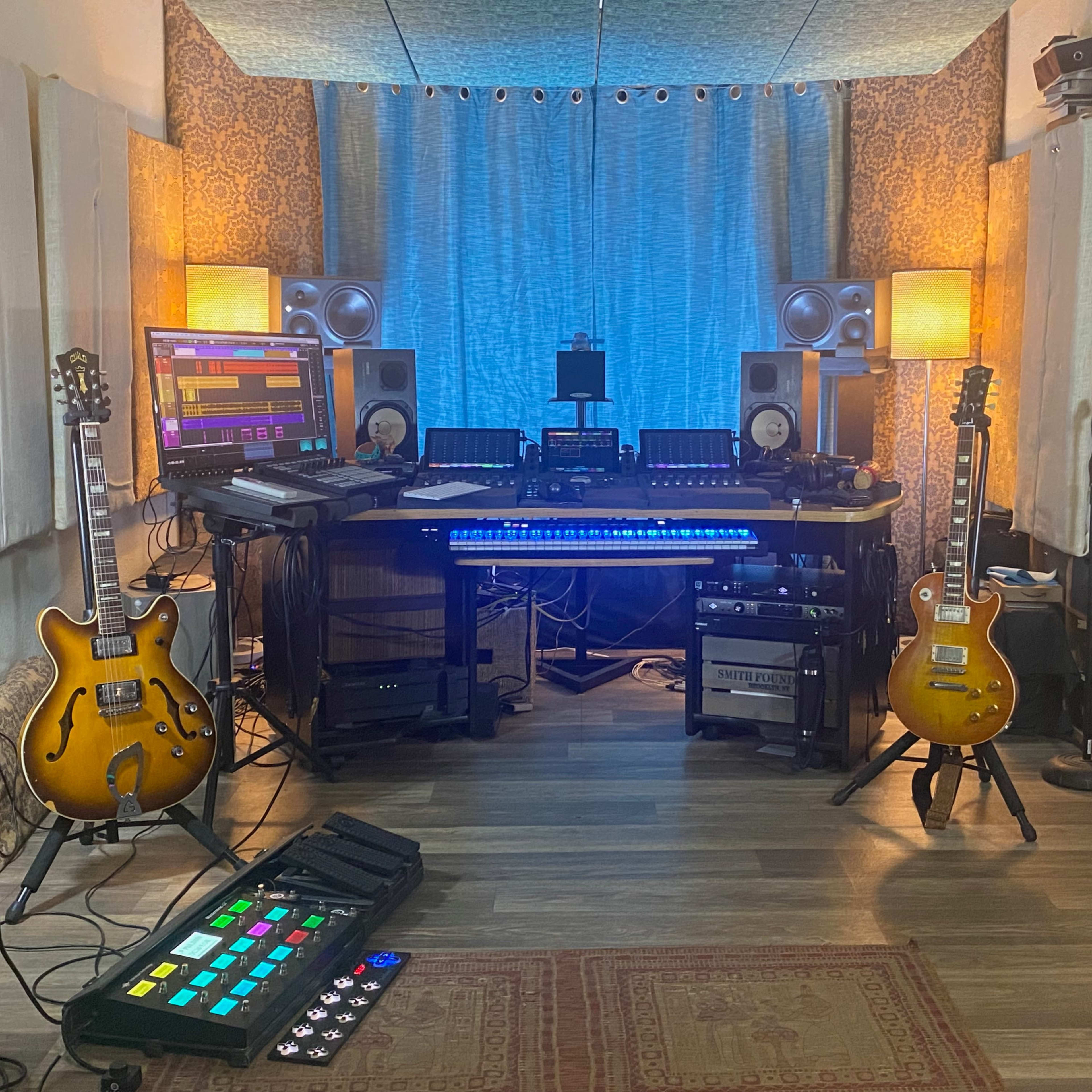 A music studio setup featuring two electric guitars, multiple monitors, audio equipment, and a MIDI controller, all arranged in a cozy room with soundproofing panels.