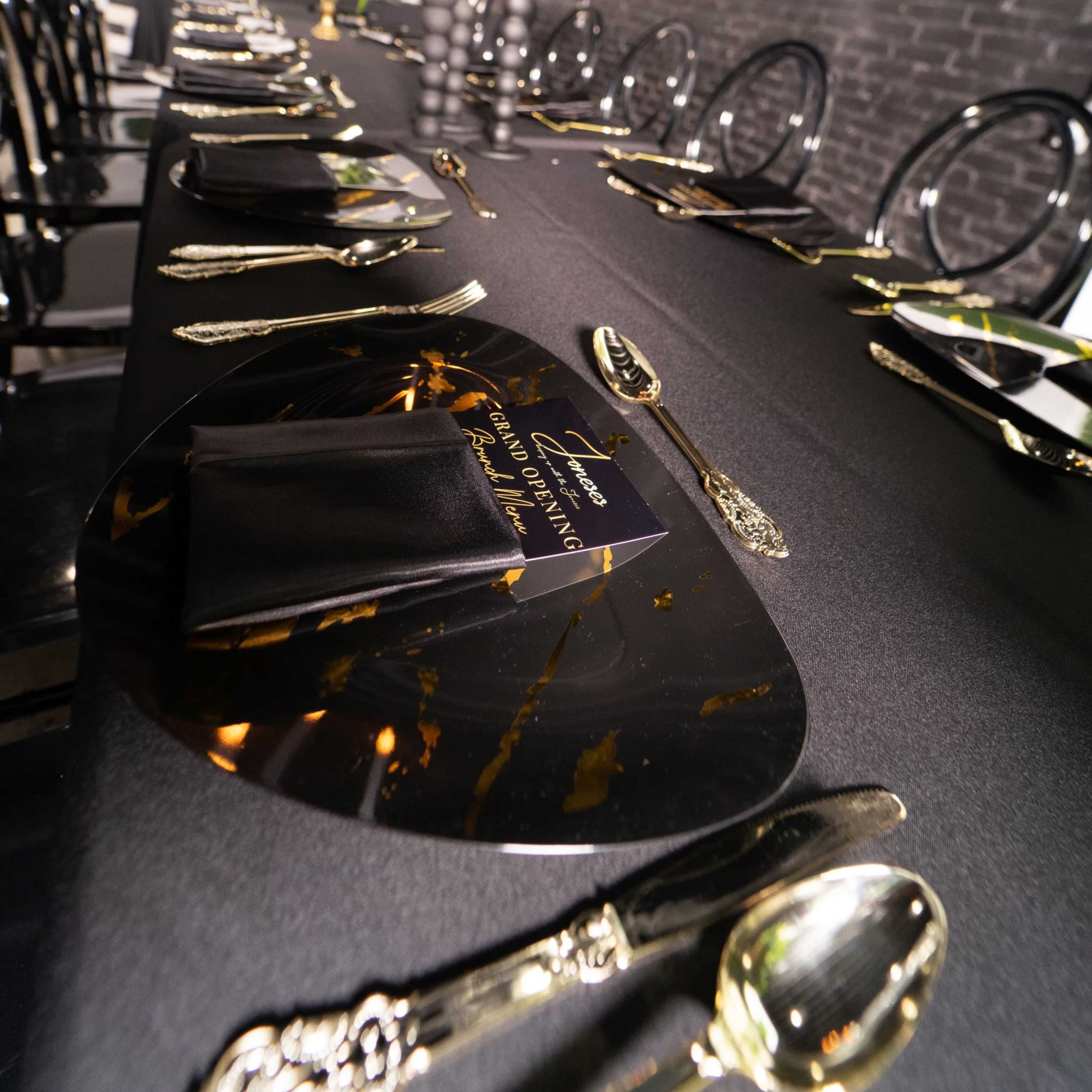 A long dining table is set with black and gold tableware, black napkins, and decorative candles, against a backdrop of dark brick walls.
