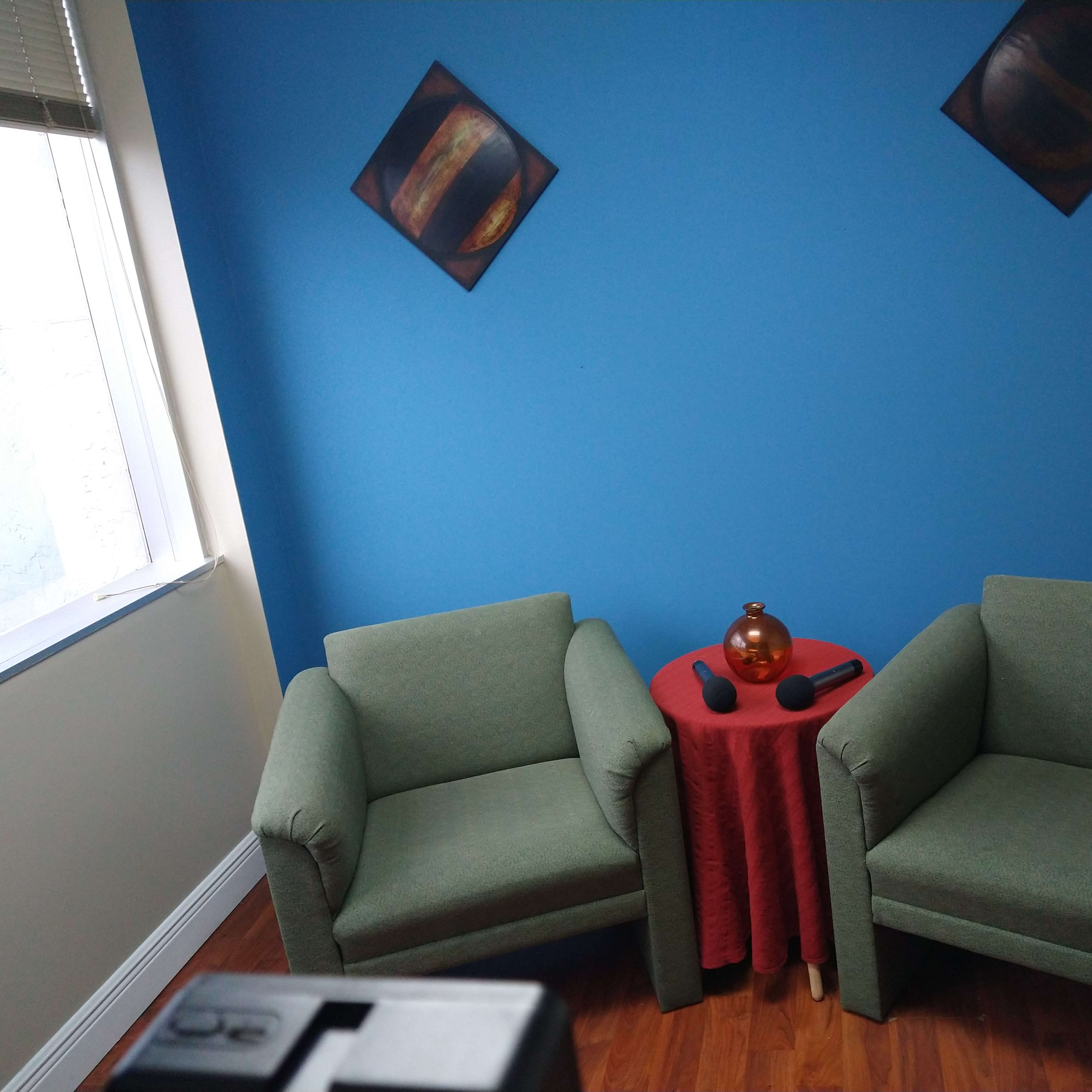 Two green armchairs positioned around a small red table with a brown vase and two microphones, against a blue wall.