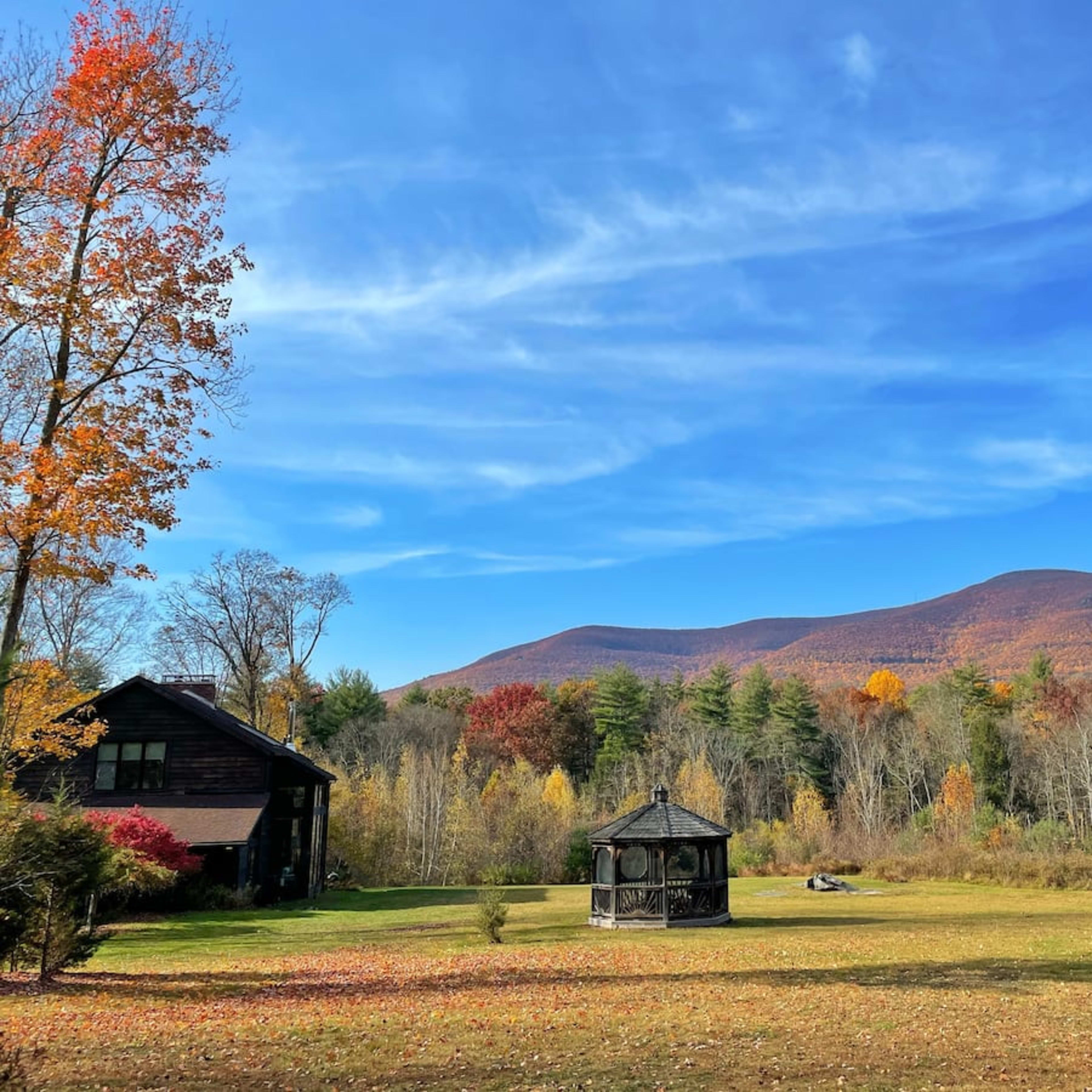 A black house with a gazebo is set against a backdrop of colorful autumn trees and mountains under a clear blue sky.
