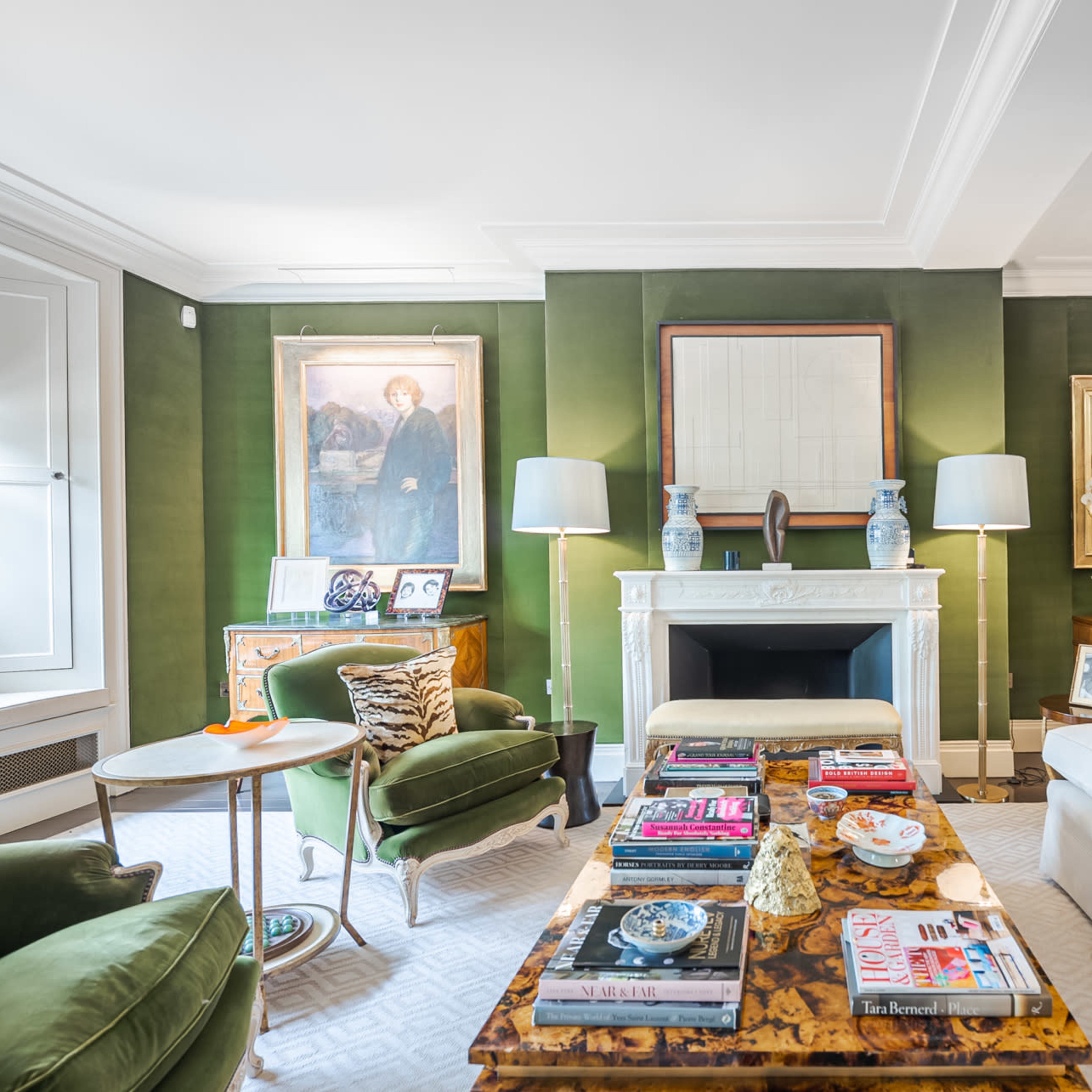 The living room features a green wall, a white fireplace with decorative art above it, and a mix of modern and antique furniture arranged around a coffee table stacked with books and decorative items.